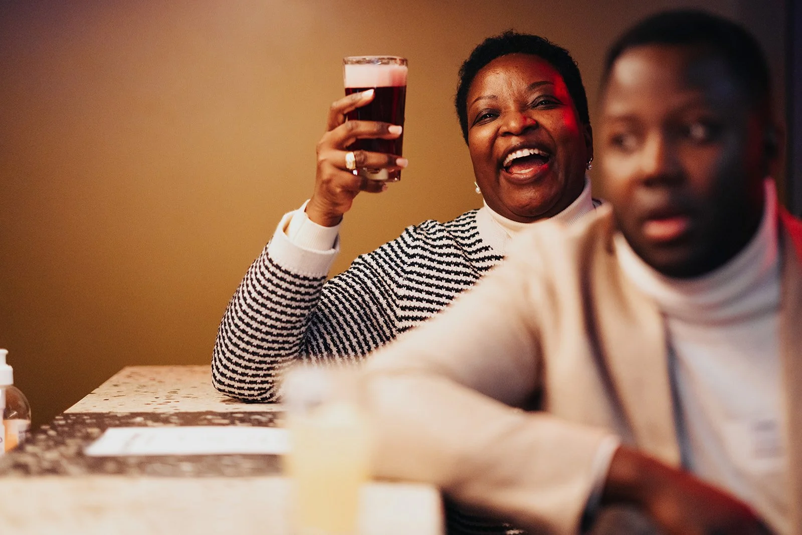 Woman holding up a glass of dark beer and smiling, sitting at a table with another person in the foreground.