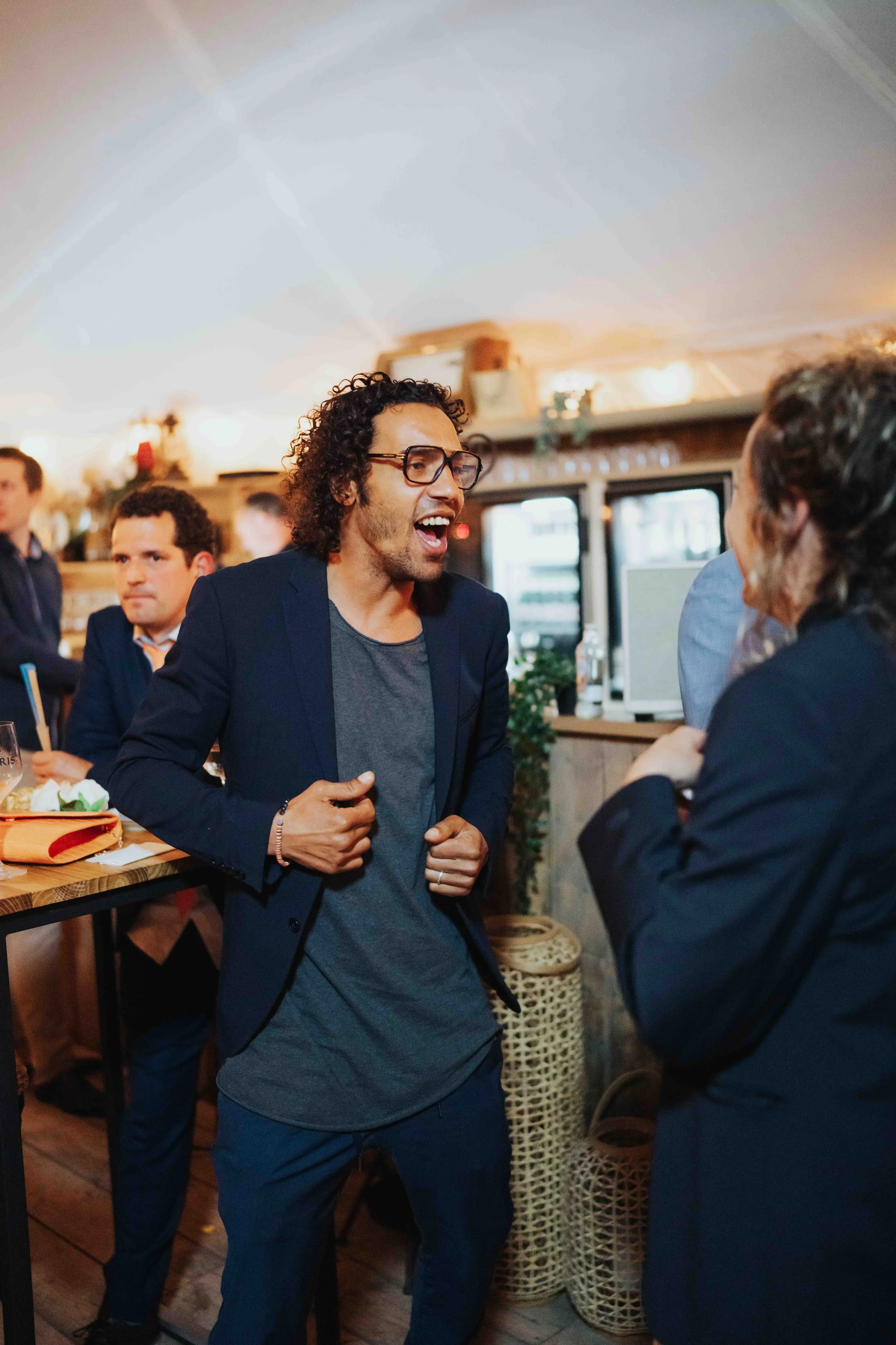 A man with curly hair, glasses, and a dark blazer is animatedly talking to a woman or dancing in a party tent or bar, with other guests seated nearby. The setting is cozy with warm lighting and wooden decor.