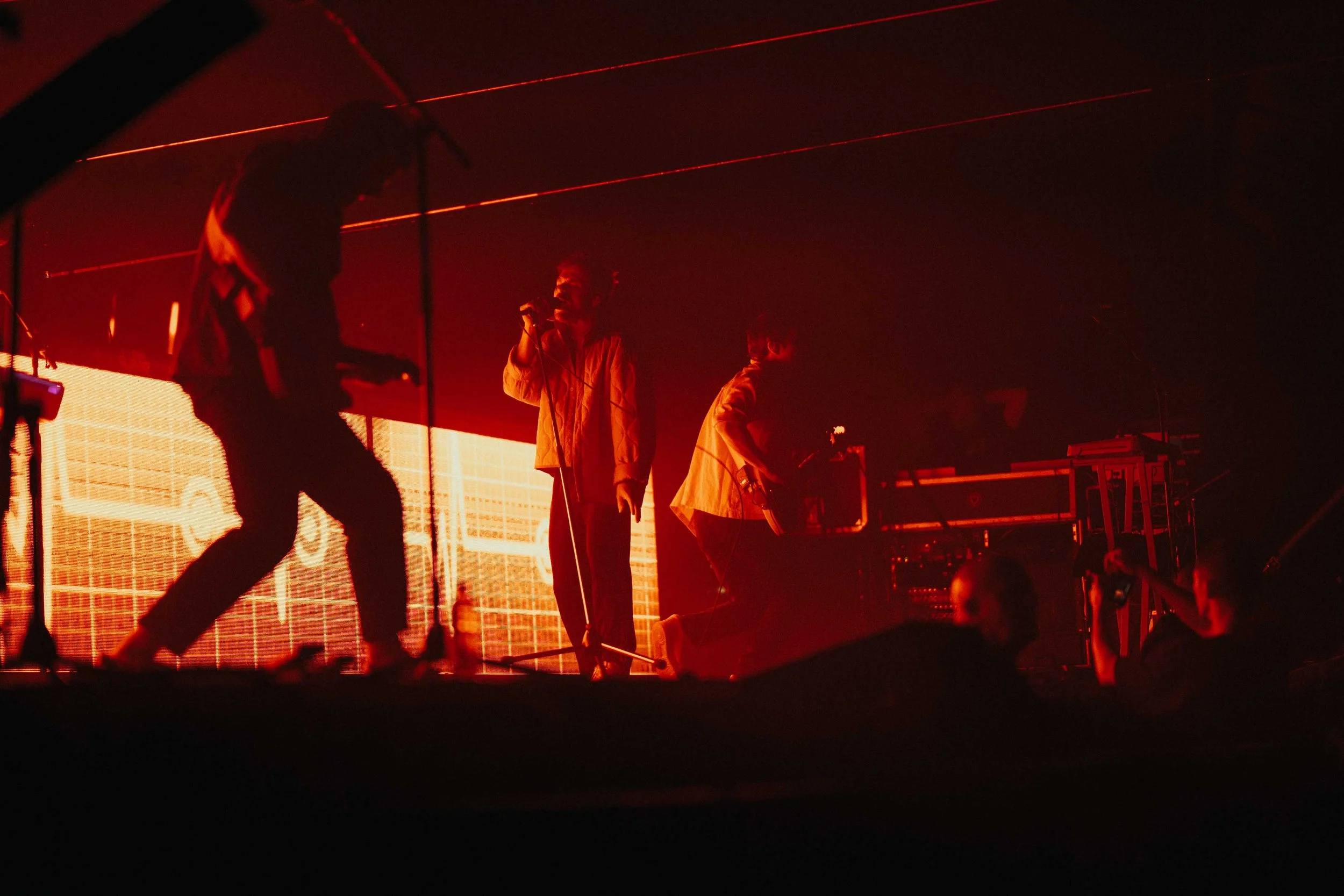 Silhouettes of three members of the band Enter Shikari performing on a stage with red lighting and a large digital screen in the background.
