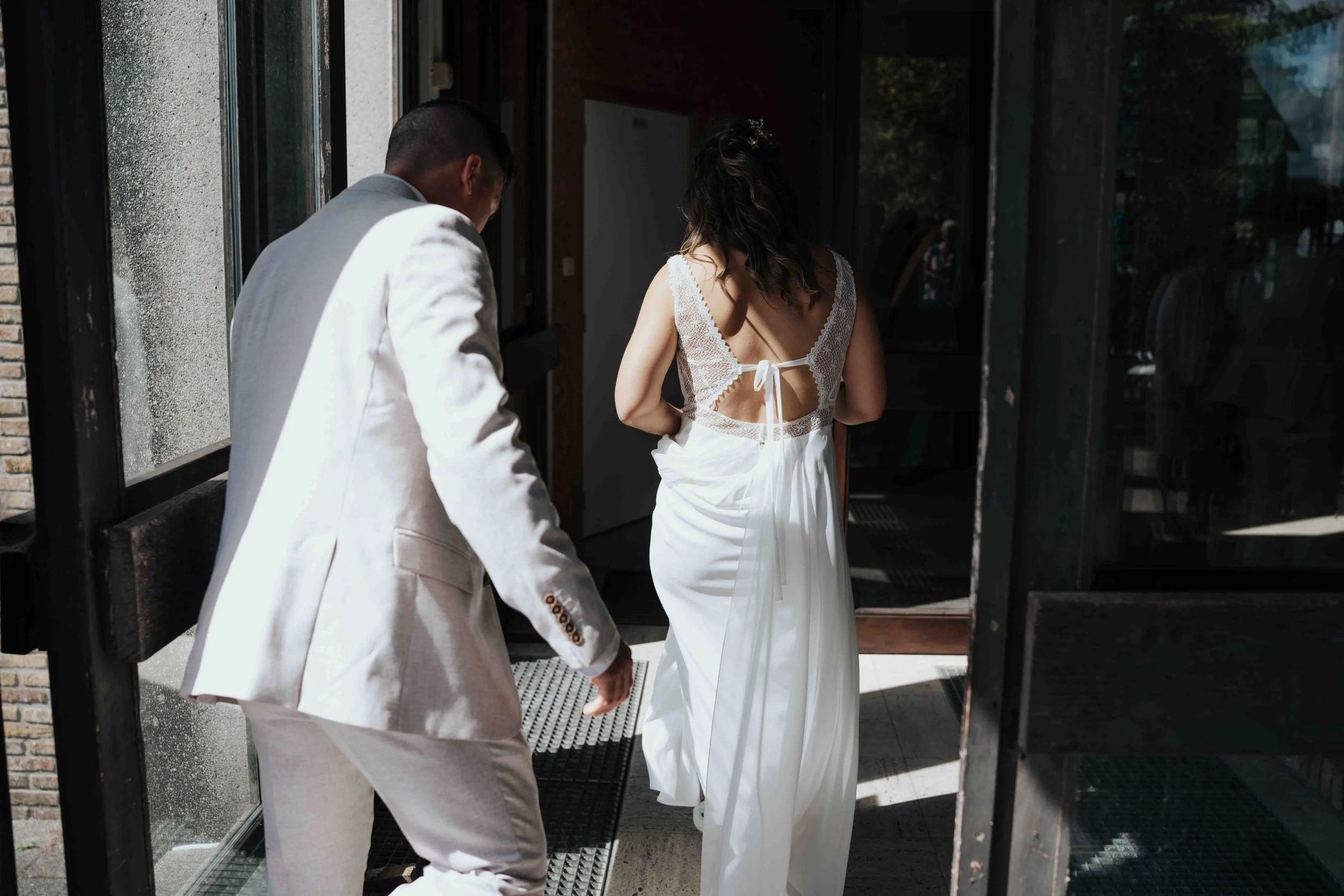 A man in a light-colored suit and a woman in a white dress are walking out of a building through a glass door, seen from behind.