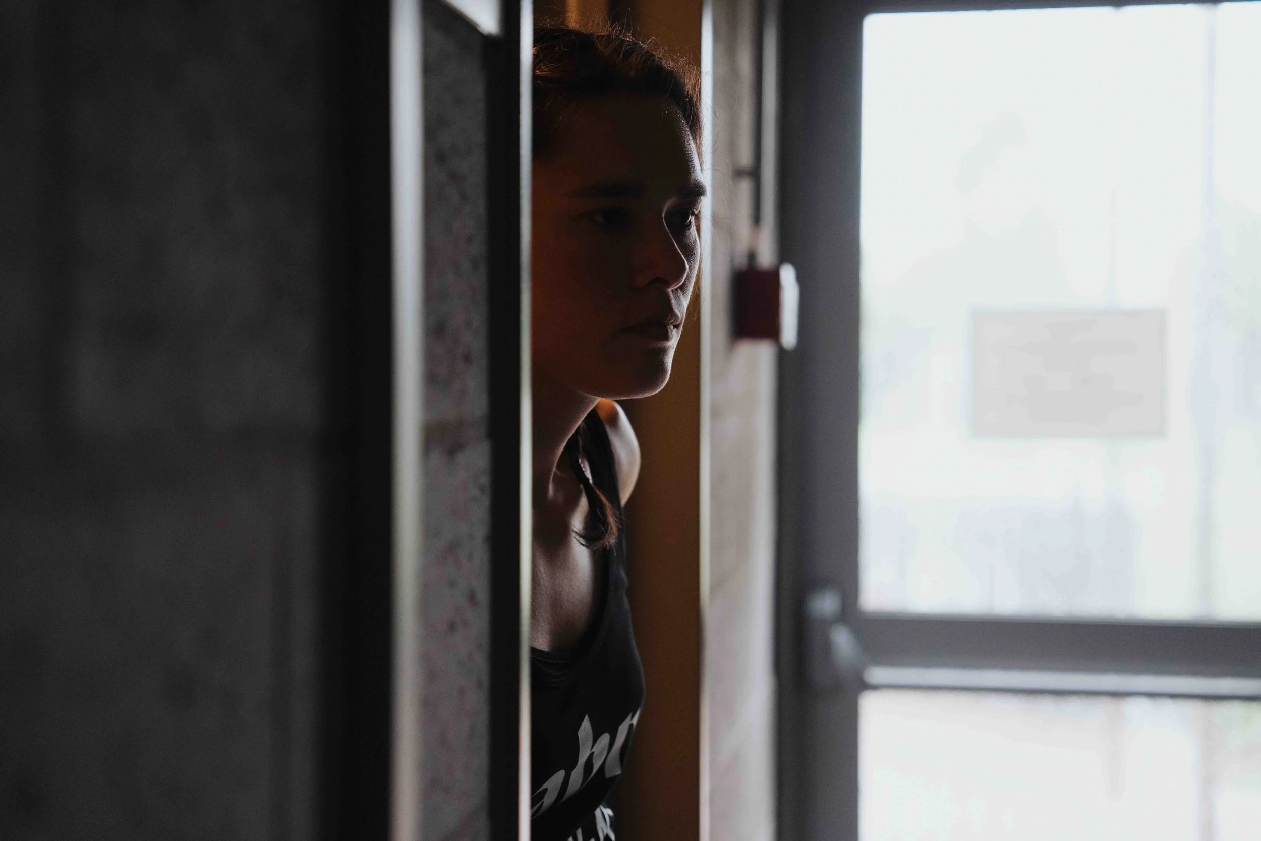 A woman is peeking out from behind a dark wall, looking toward a bright door with a window.