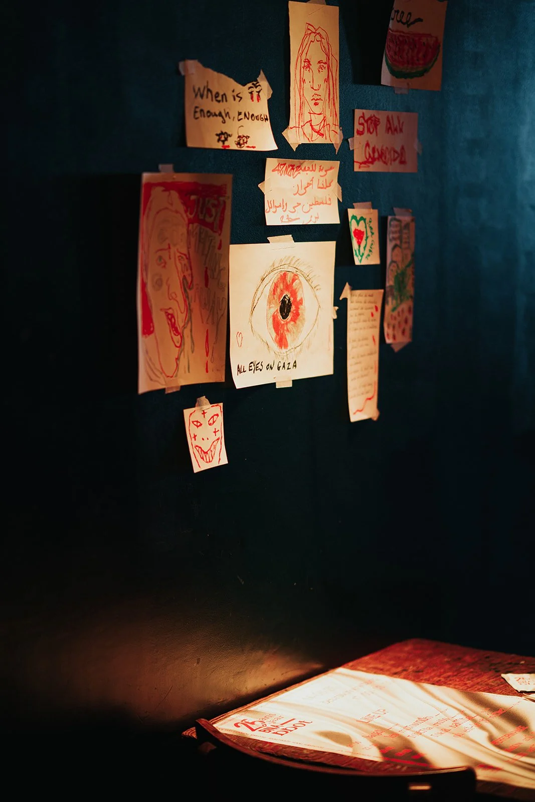 A wall with various hand-drawn notes and pictures taped to it, including sketches of faces, a large red eye with the words 'ALL EYES ON GAZA,' and messages like 'Stop All Crimes.' A book is open on a desk below with sunlight casting shadows on it.