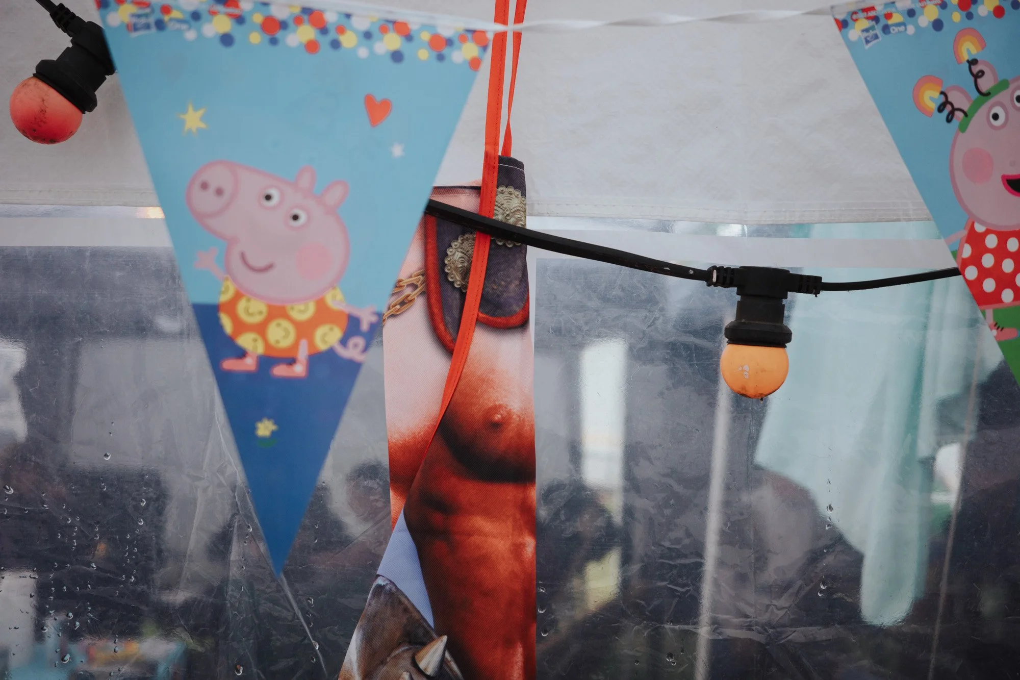 Close-up of blue Peppa Pig-themed party banners with purple, yellow, and red details, hanging on a string with a black and orange string light visible and a male nipple apron, with a blurred background and a plastic sheet.