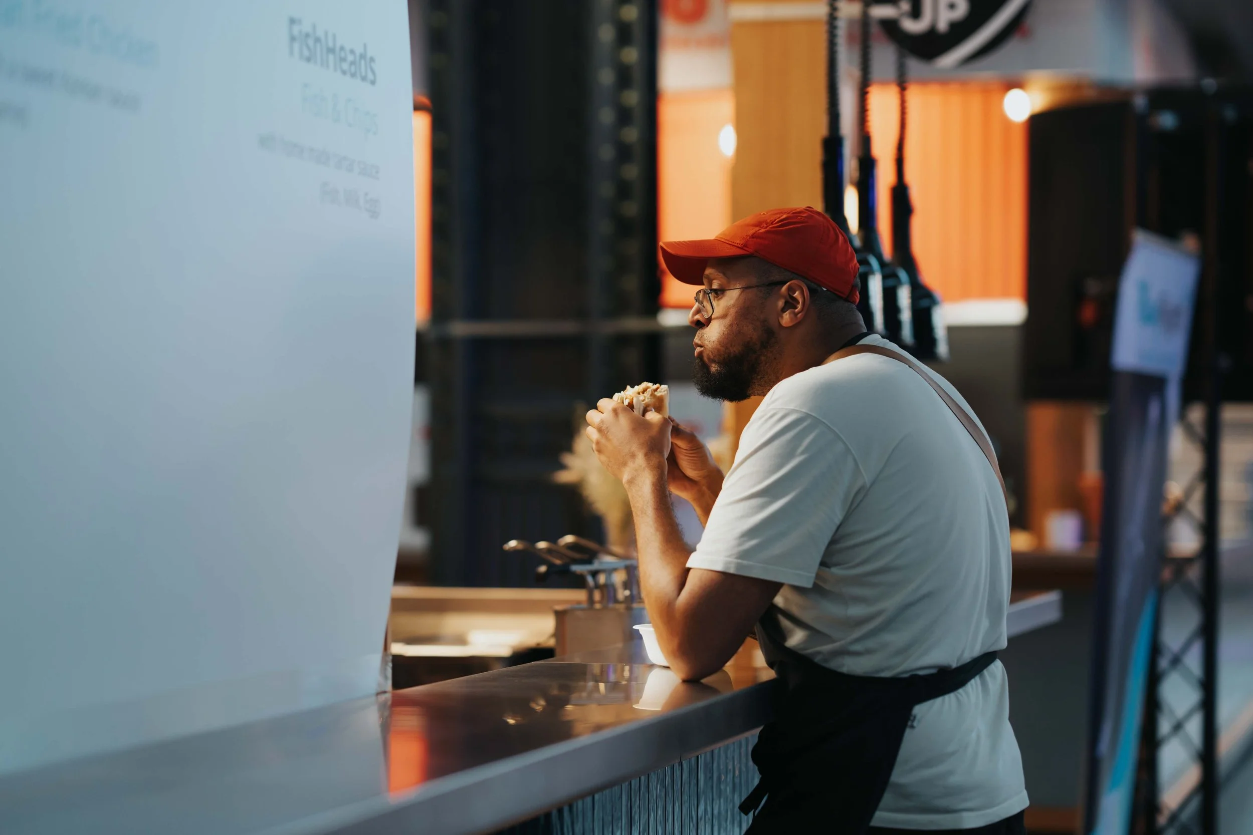 A man with glasses, a red cap, and a white shirt stands at a food stand, eating a sandwich or burger, with a dark background and warm lighting.