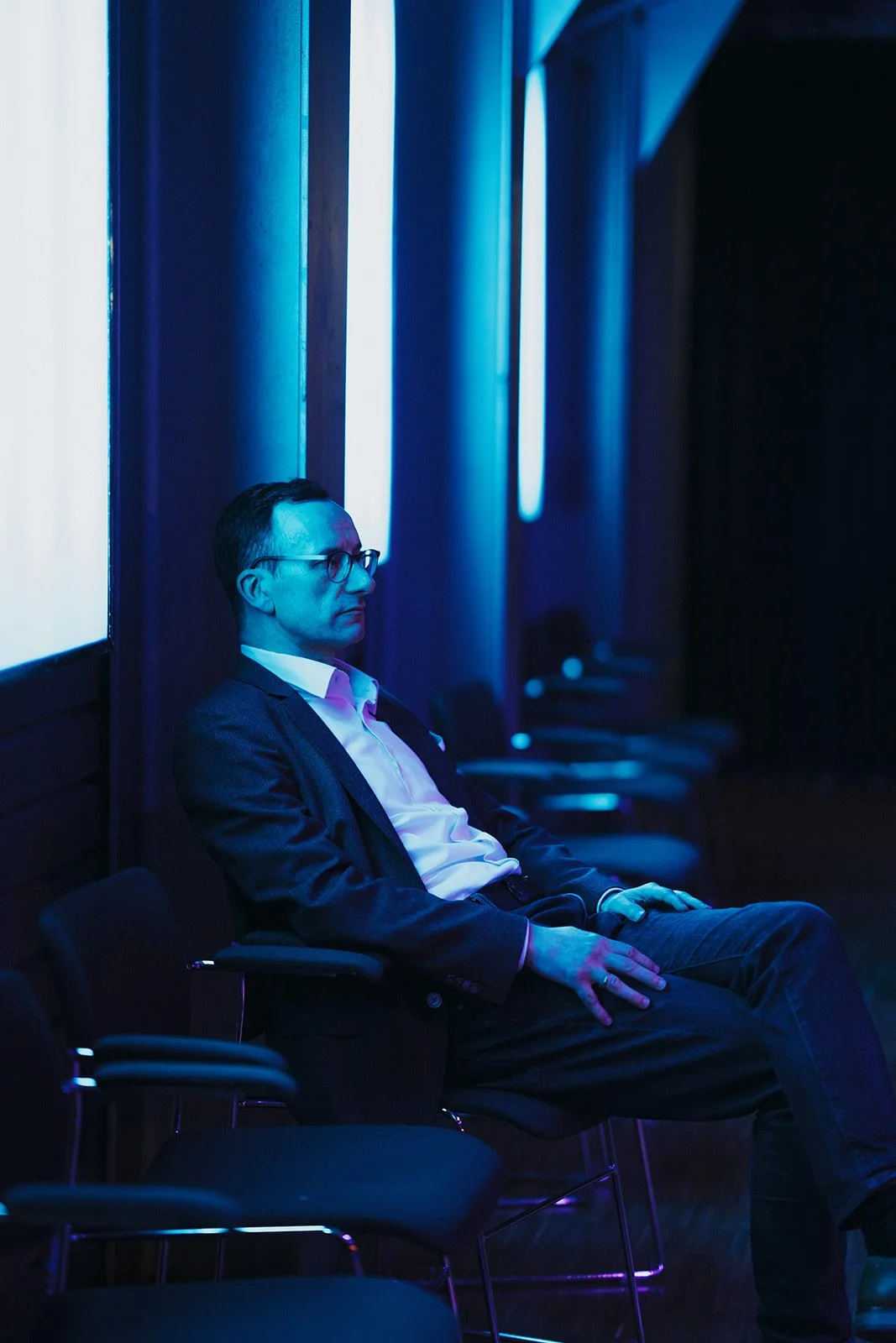 A man in business attire and glasses is sitting on a chair in a dimly lit room with blue lighting, looking to the left.