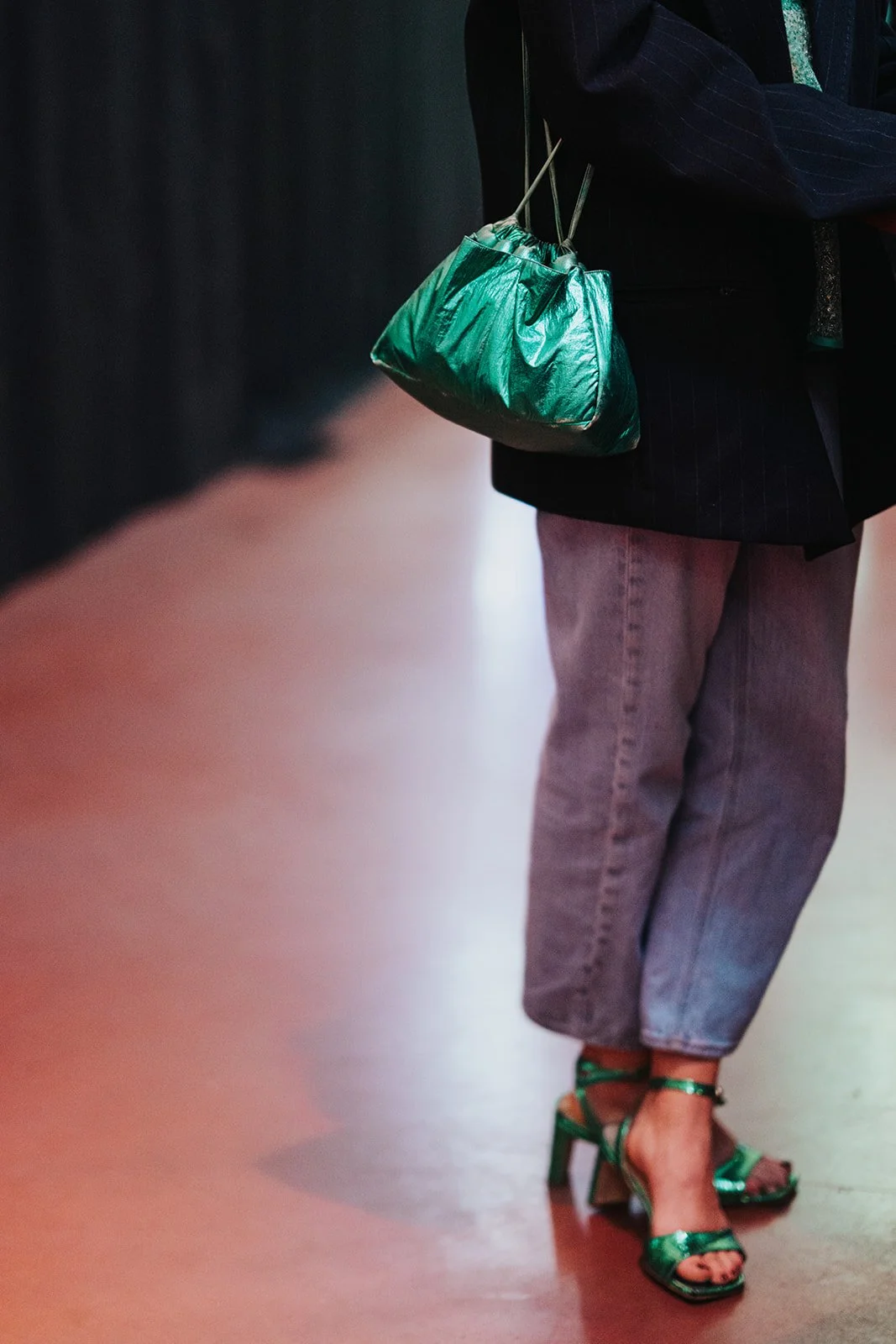 Close-up of a person holding a shiny green pouch, wearing gray jeans and green high-heeled sandals, with part of a dark blazer visible.
