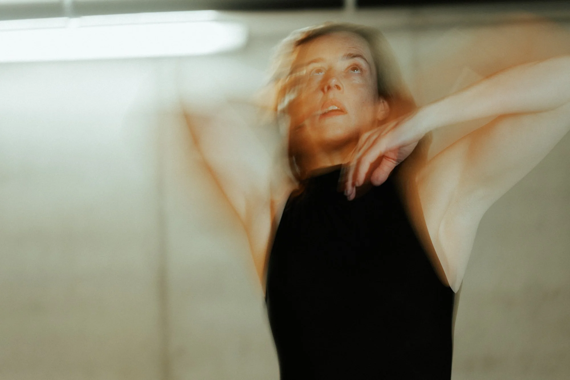 A woman stretching with her arms raised and her hand touching her neck, wearing a black sleeveless top, in a room with soft lighting.