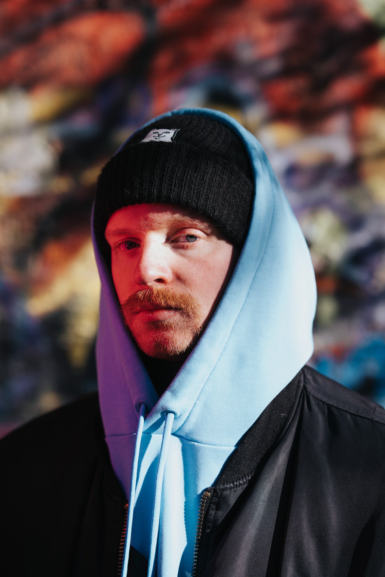 A man with light skin, blond hair, a beard, and blue eyes wearing a black beanie, a light blue hoodie, and a black jacket. The background is blurred with colorful abstract shapes.