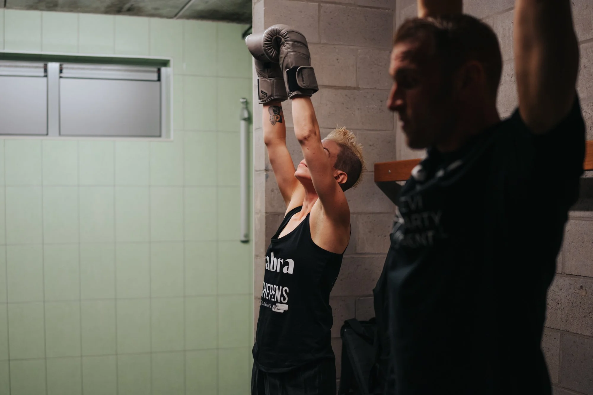A woman with short blonde hair lifting boxing gloves above her head in a locker room, with a man leaning against the wall in the foreground, both wearing workout clothing.
