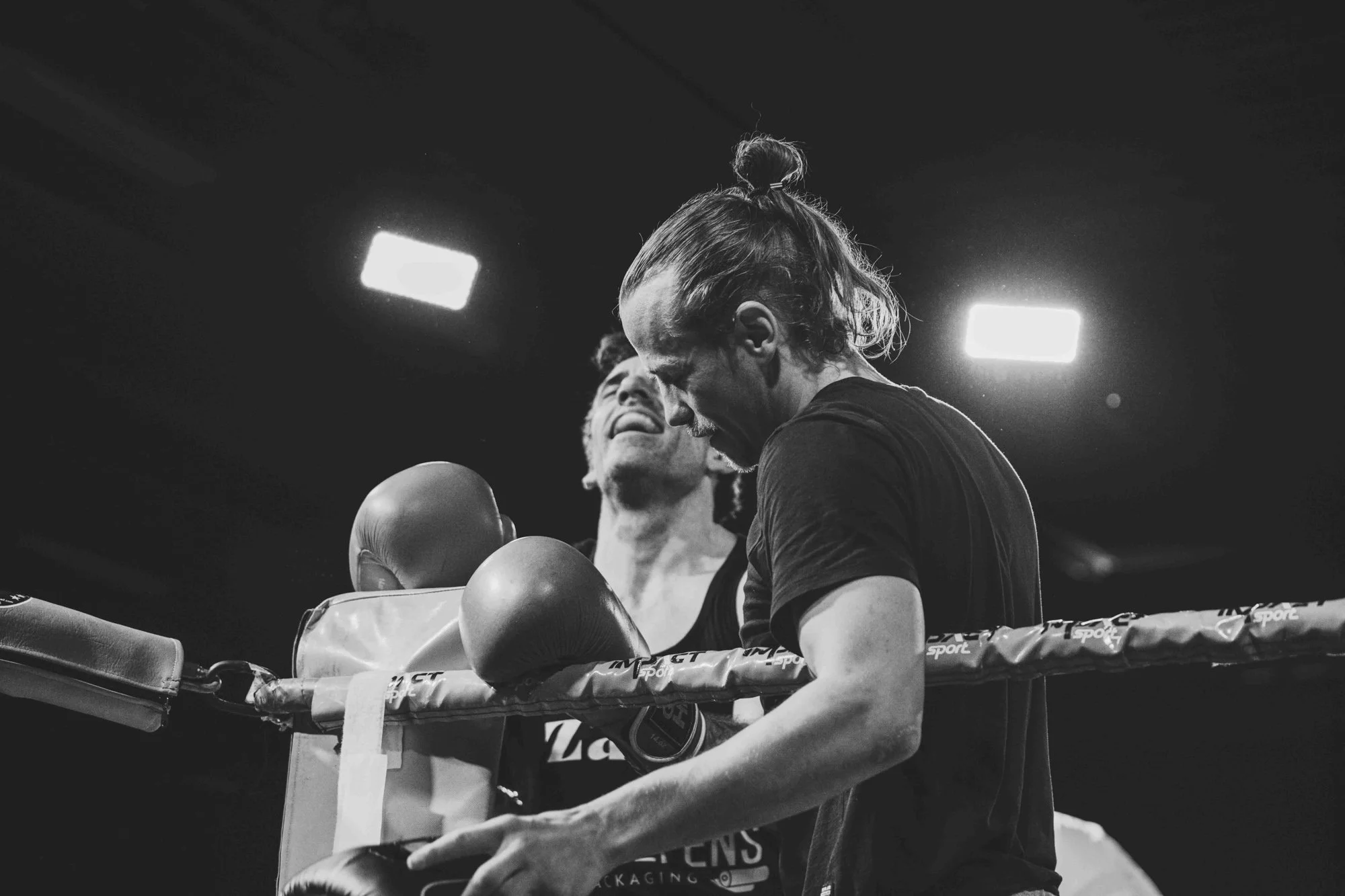 Two men in a boxing ring, one wearing boxing gloves and the other is Junior Bauwens with his head bowed, smiling, with bright lights overhead, black and white photo.