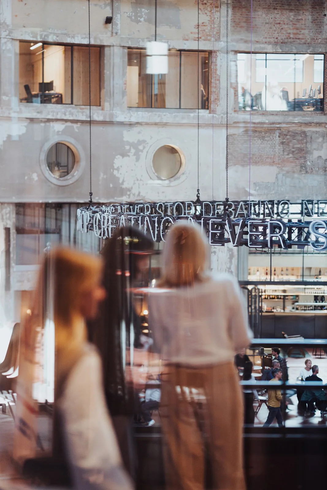 People inside a modern building with large windows, looking at mannequins and a neon sign that reads 'NEVER STOP DREAMING, NEVER STOP CREATING' at Wintercircus Ghent.