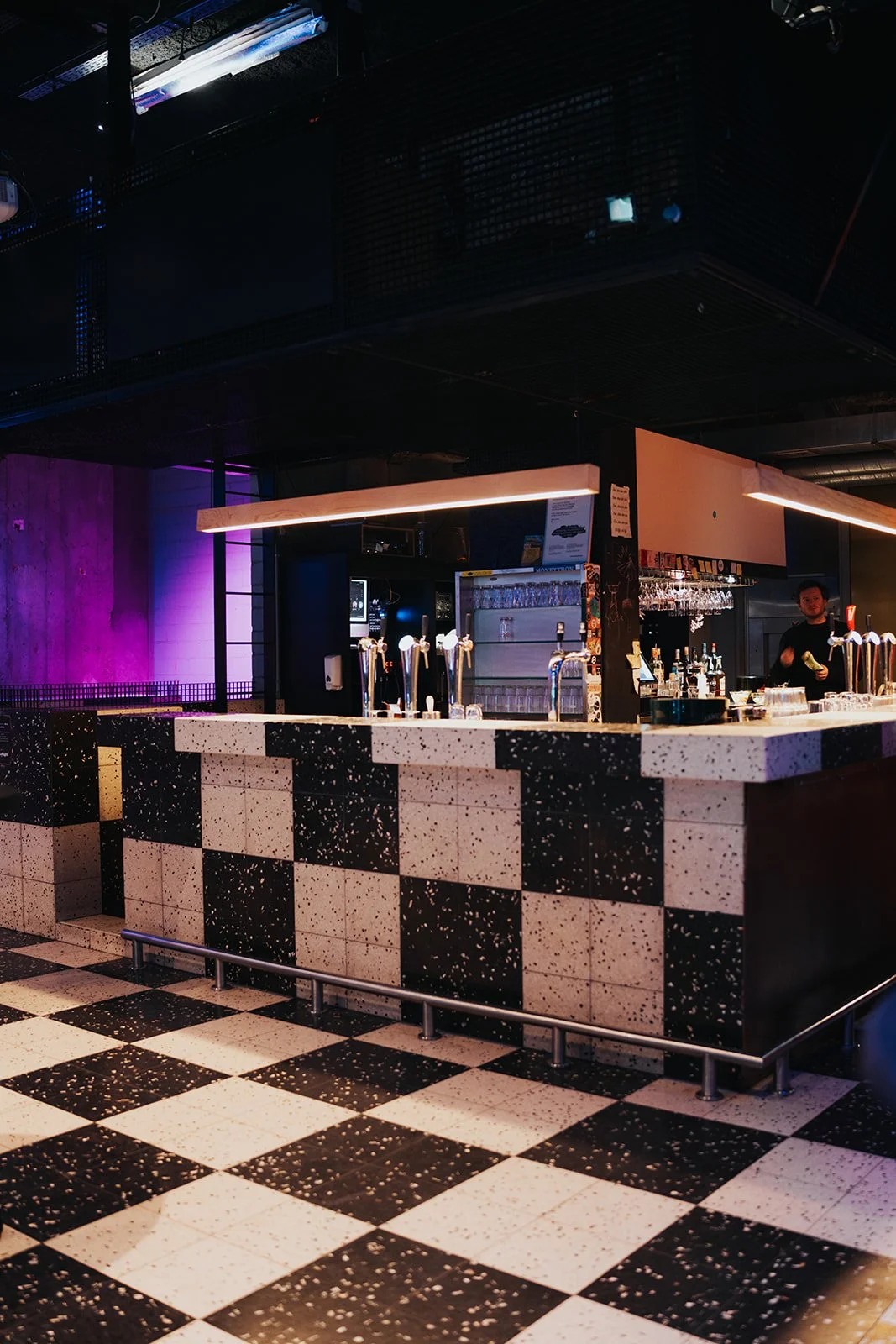 A bar with a black and white checkered floor and a bar counter with taps for draft beer. The background includes shelves with glasses, bottles, and a bartender preparing drinks. The lighting features colorful, ambient lighting with purple and blue hu