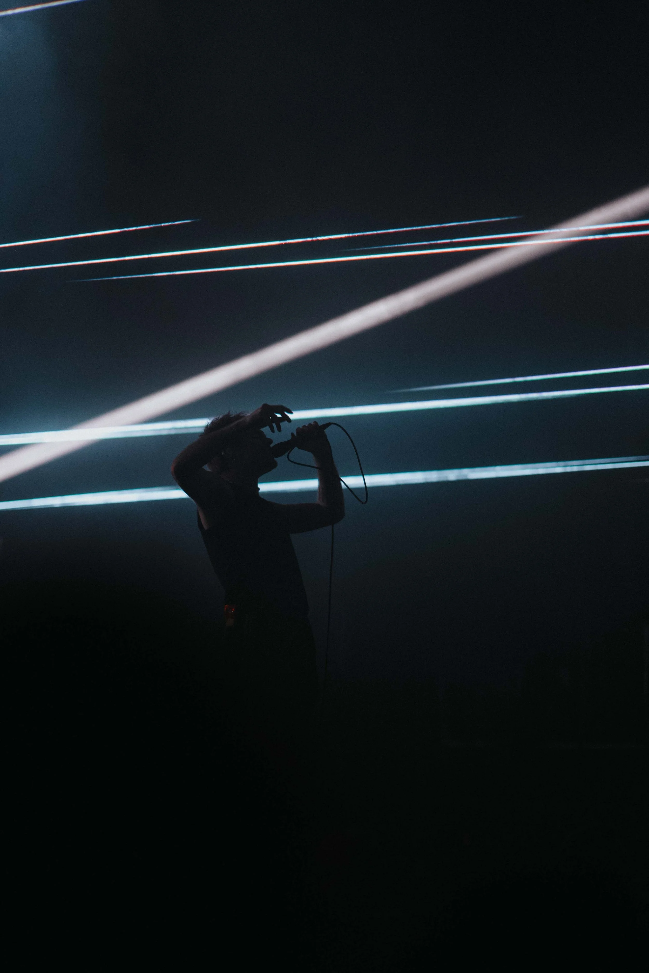 Silhouette of Rou Reynolds of Enter Shikari singing into a microphone on stage with colorful light beams and laser effects in the background.