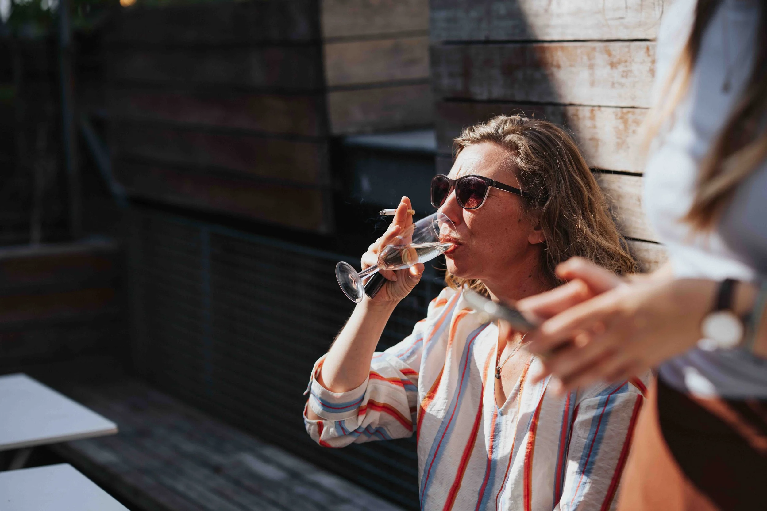 Woman with sunglasses drinking champagne outdoors, sitting against a wooden wall, holding a cigarette in her right hand, while another person beside her holds a phone.