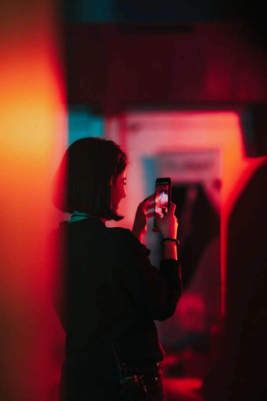 A woman with short dark hair is standing and taking a photo with her smartphone in a dimly lit room with red and blue lighting.