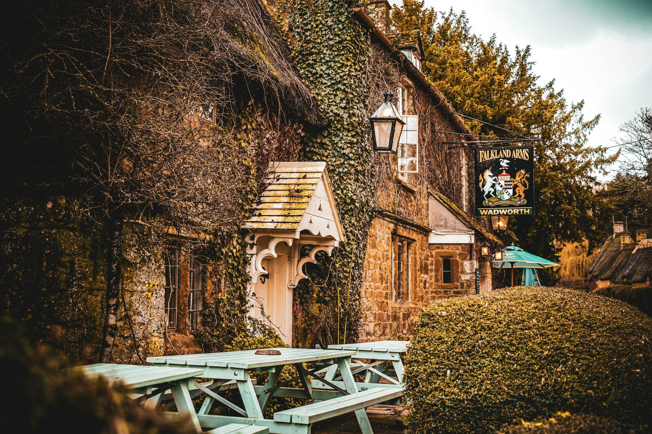 An exterior scene of the Falkland Arms pub in Wadhurst with stone building walls, ivy, and outdoor seating including green picnic tables, near bushes and trees.
