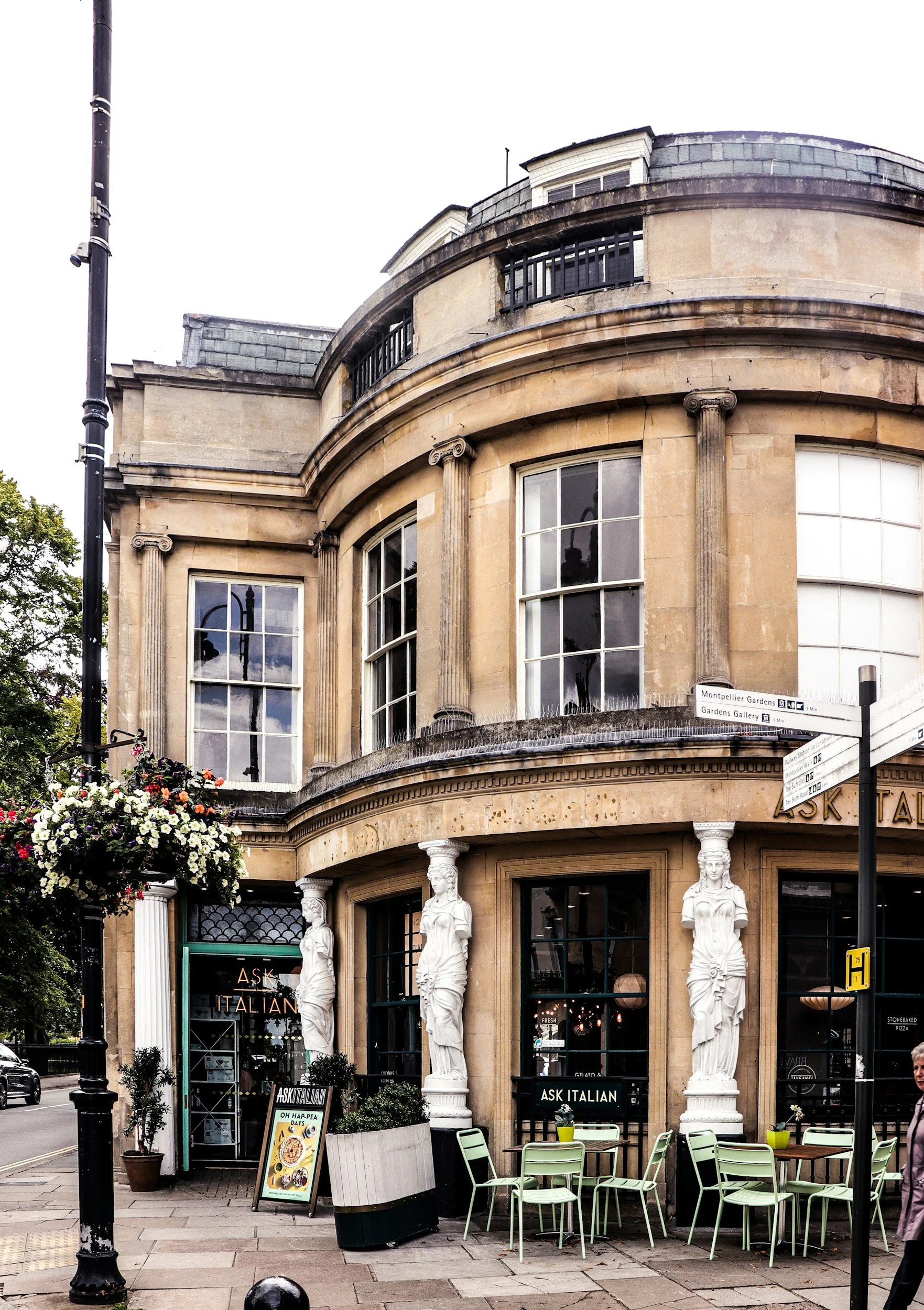 A historic building with classical architecture features, including columns and large windows, housing a restaurant called 'Ask Italian', with outdoor seating and decorative statues at the entrance.
