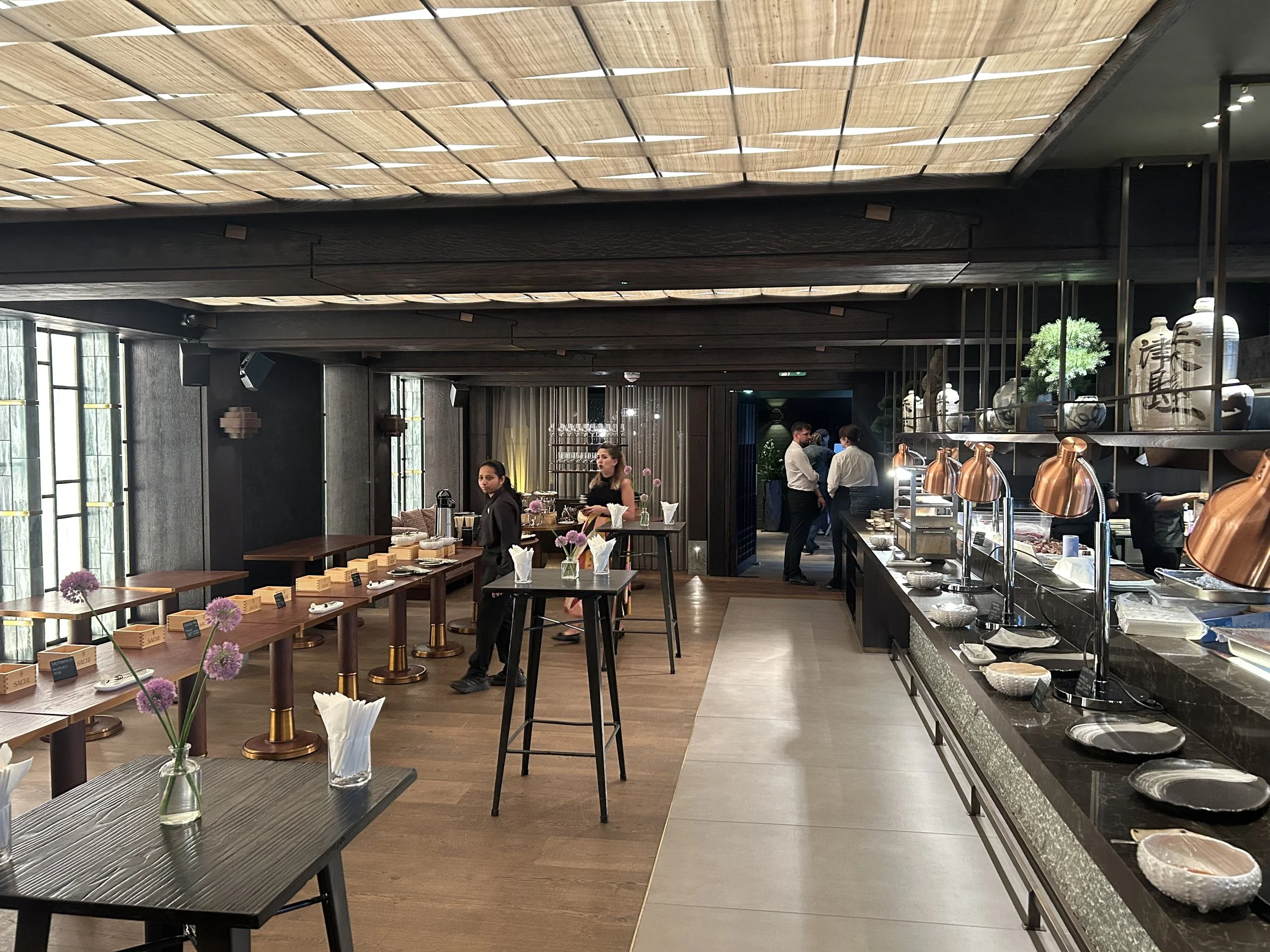 Interior of a modern restaurant with a buffet station, staff preparing food, and tables with flowers and tableware.