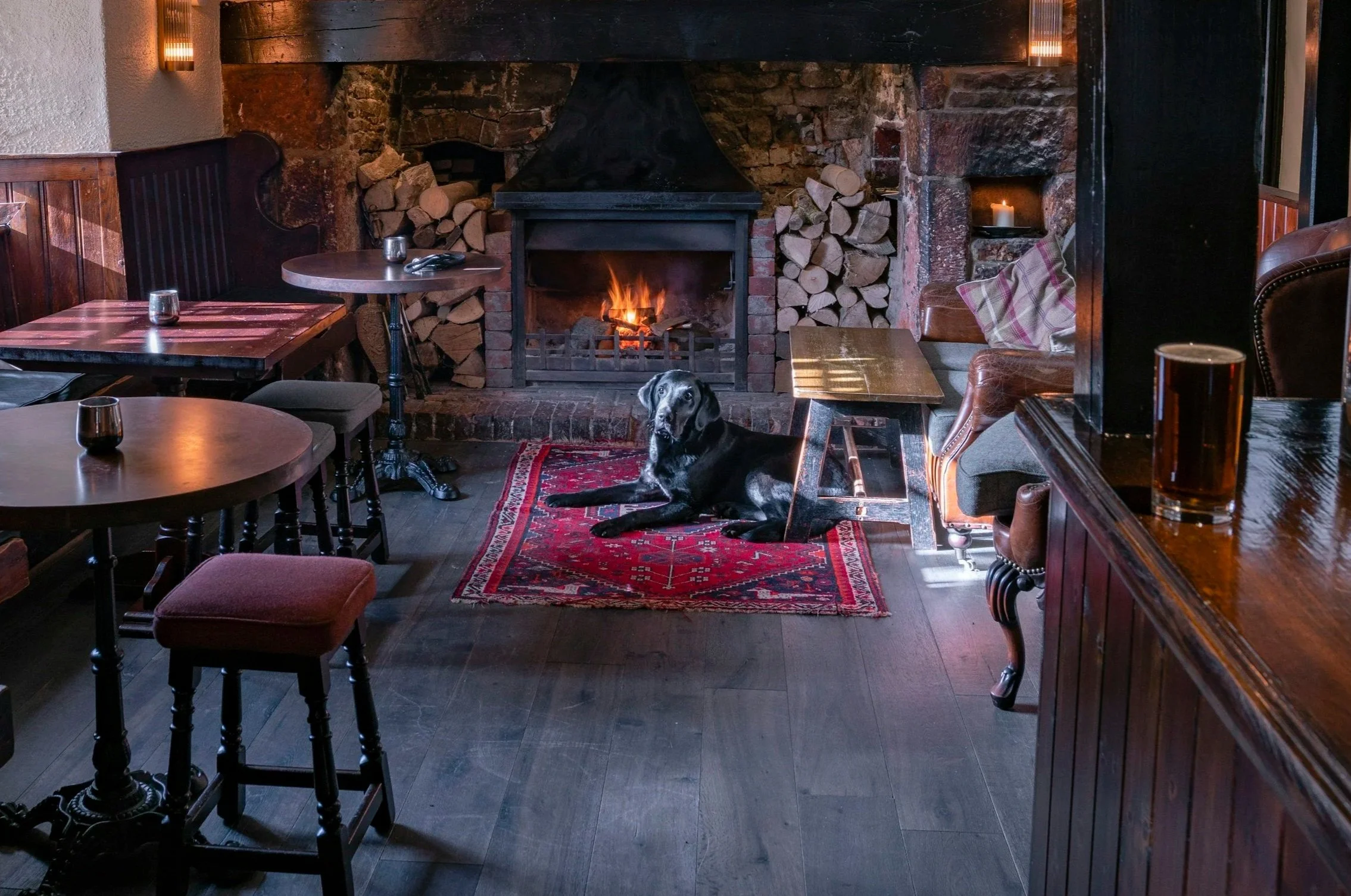 A cozy room featuring a lit fireplace with stacked logs on both sides, a large black dog lying on a small red rug, with wooden tables and chairs, a candle, and a candle holder creating a warm ambiance.