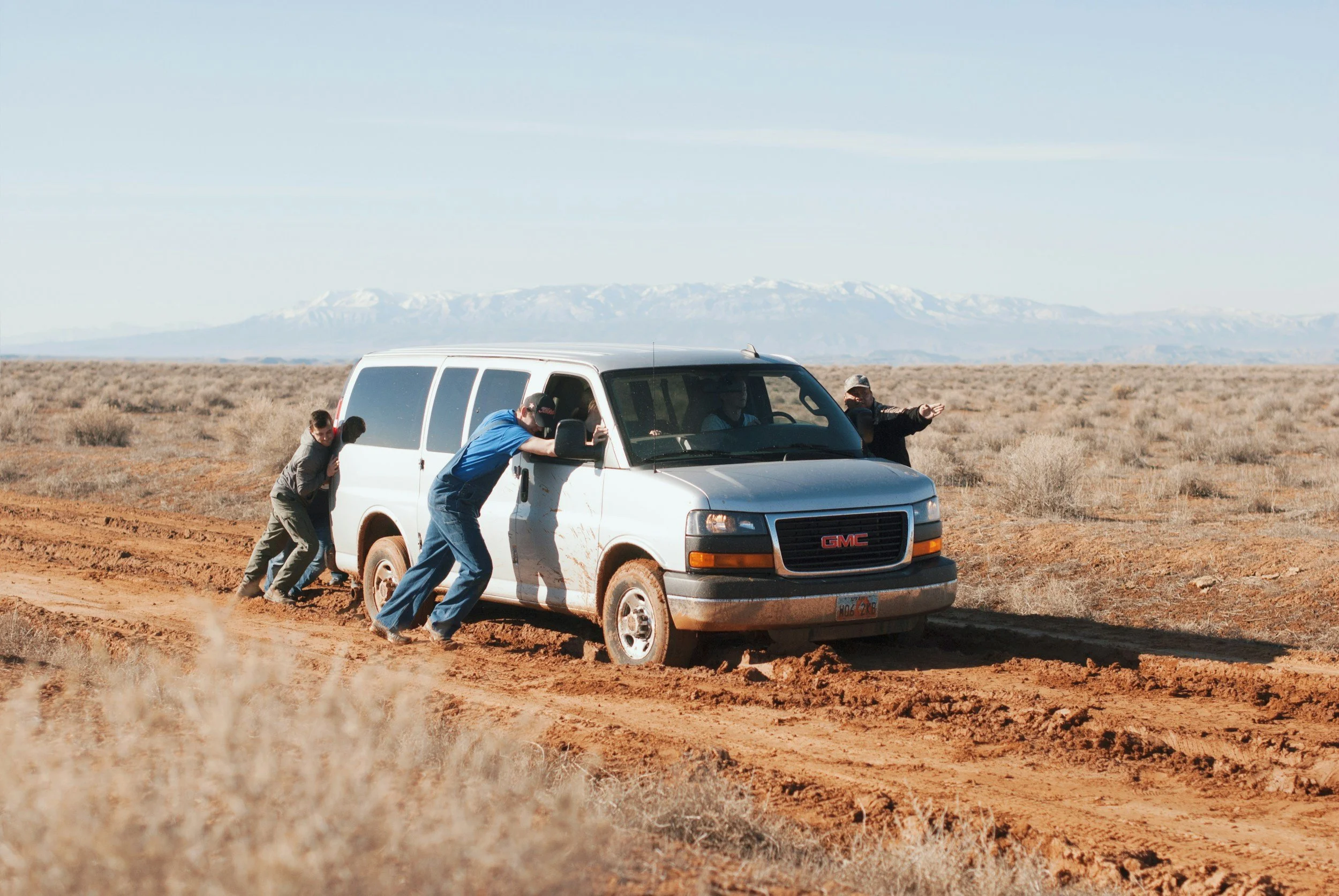 Four people attempting to push a stuck white van with a GMC logo on a dirt road in a dry, open landscape with mountains in the background.