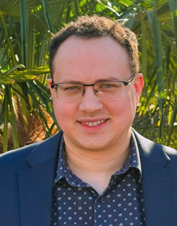 A man with short curly hair, glasses, and a friendly smile, wearing a dark blue suit jacket and a dark blue shirt with white polka dots, standing outdoors in front of tall green plants.