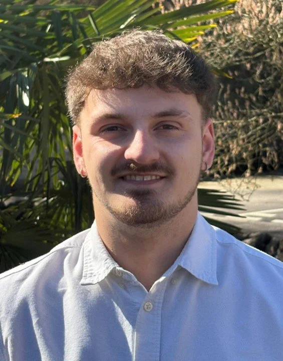 A young man with short brown hair and a beard, smiling outdoors in front of green tropical plants, wearing a white button-up shirt.