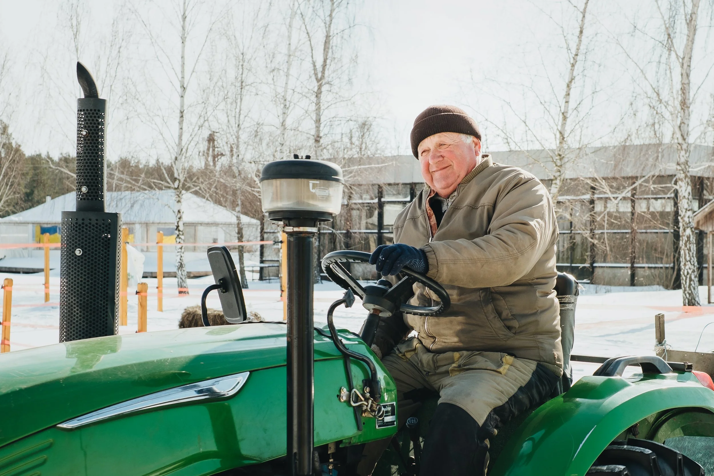 An elderly man with a brown beanie hat driving a green tractor outdoors in a winter setting, with leafless trees and snow in the background.