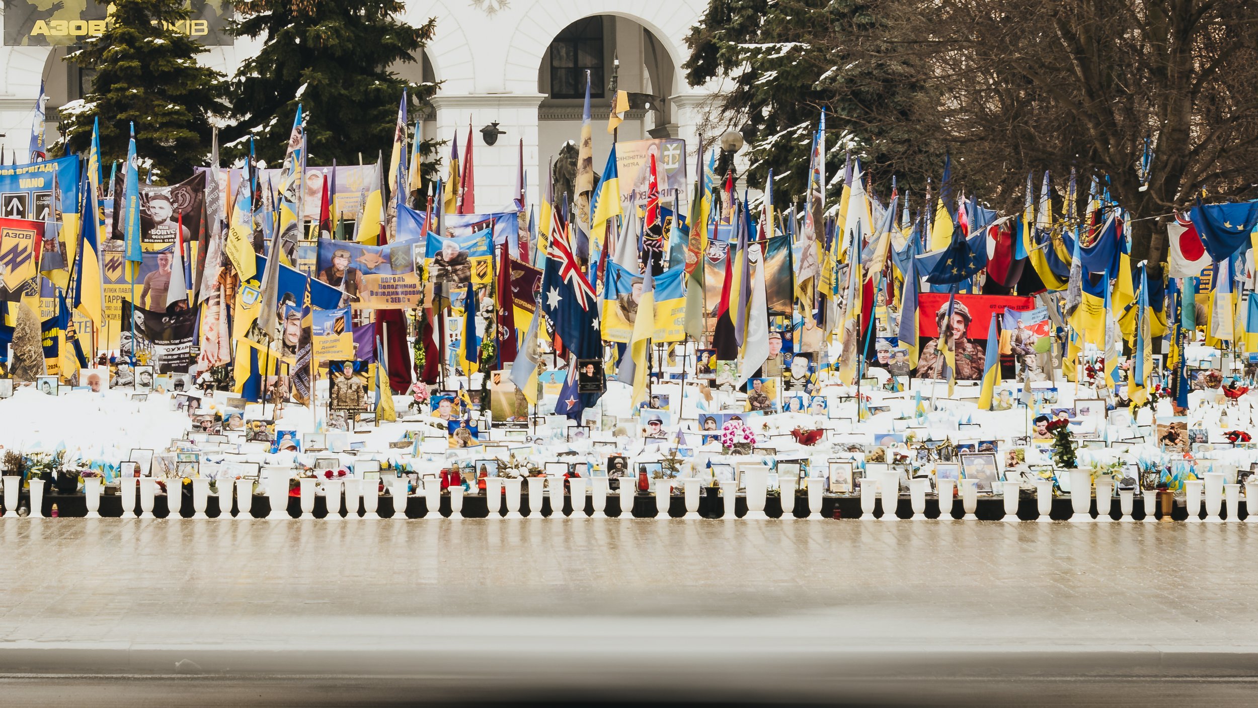 A memorial display featuring countless flags, photographs of soldiers, and memorial banners, set against a building backdrop with snow-covered trees.