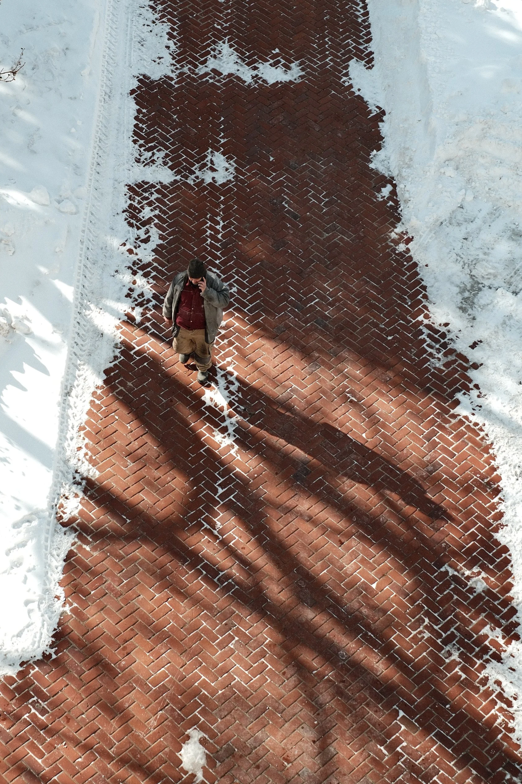 A man standing on a brick sidewalk in a snowy area, talking on his phone. The sidewalk has patches of snow and tire tracks along the edges.