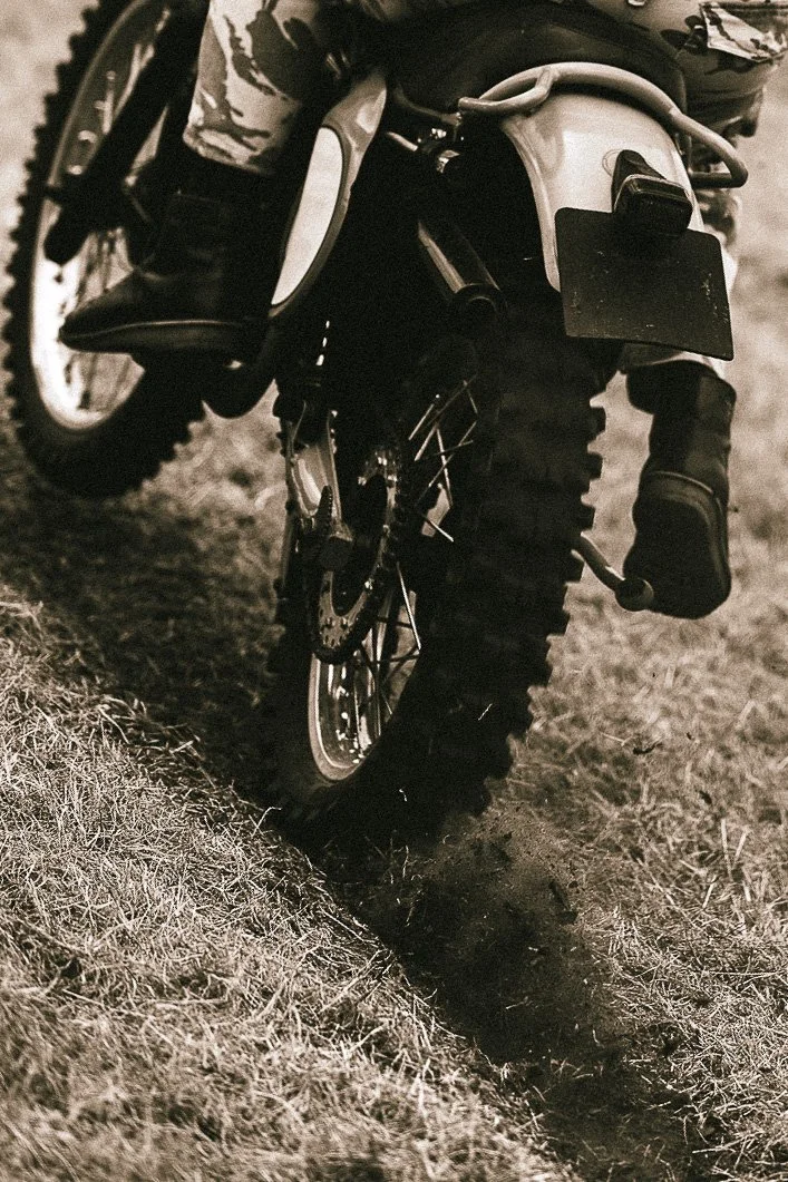 A motorcycle on uneven ground, captured in black and white, showing the rear wheel, seat, and part of the frame.