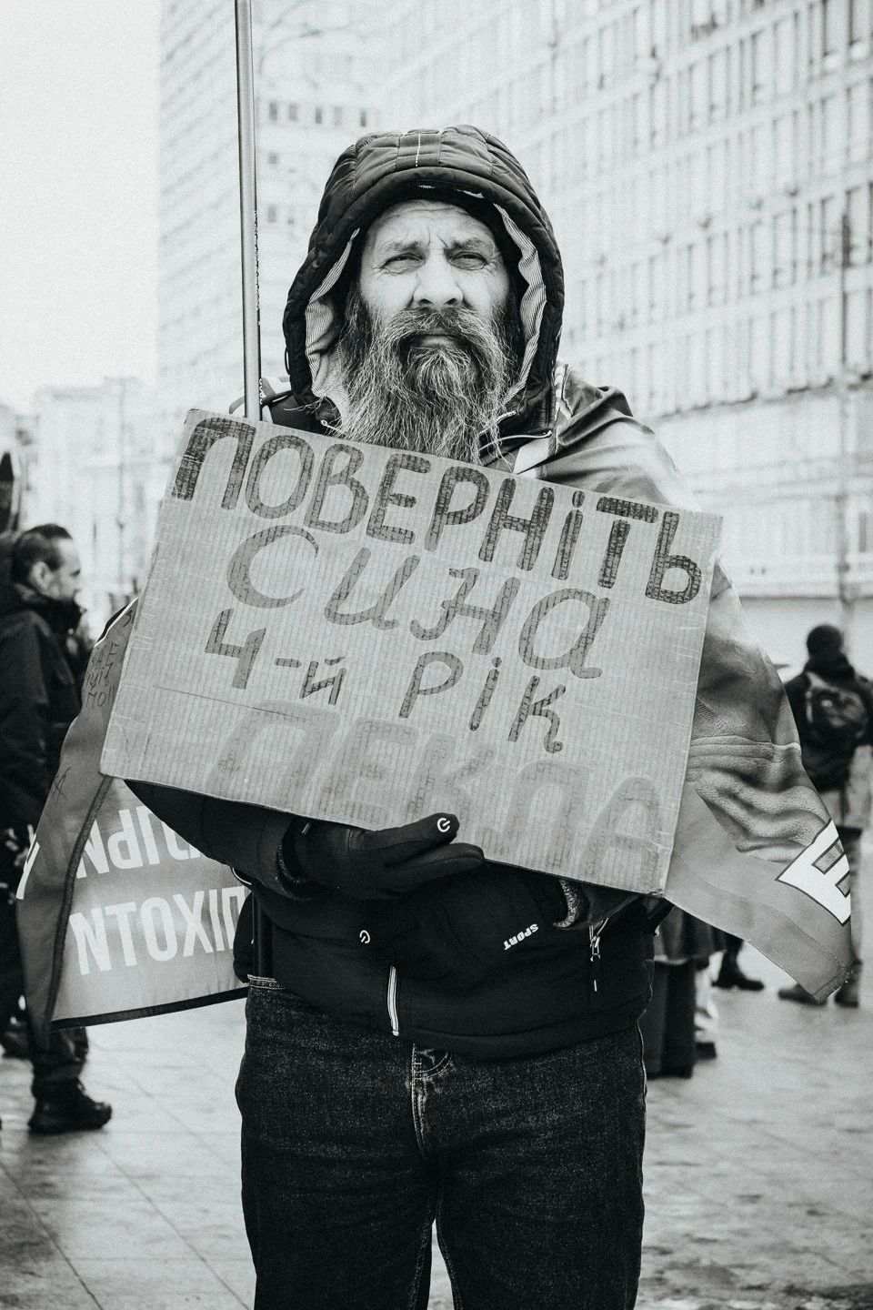 A man with a beard and a hooded jacket holding a cardboard sign written in what appears to be Cyrillic script during a street protest, with other people in the background.
