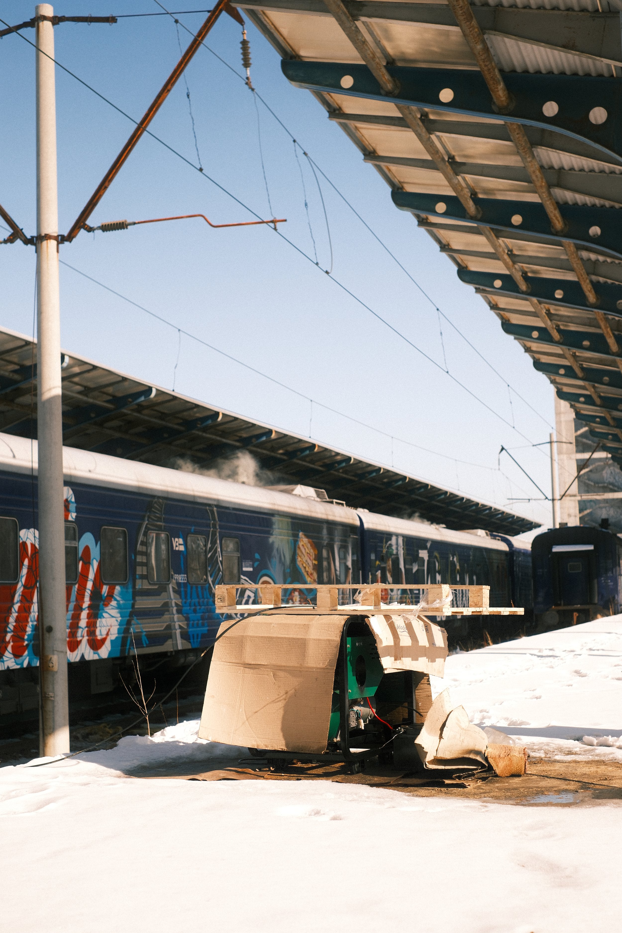 A train station platform with a train decorated with colorful graffiti art, a snow-covered ground, and construction equipment in the foreground. An overhead structure and electrical wires are also visible.