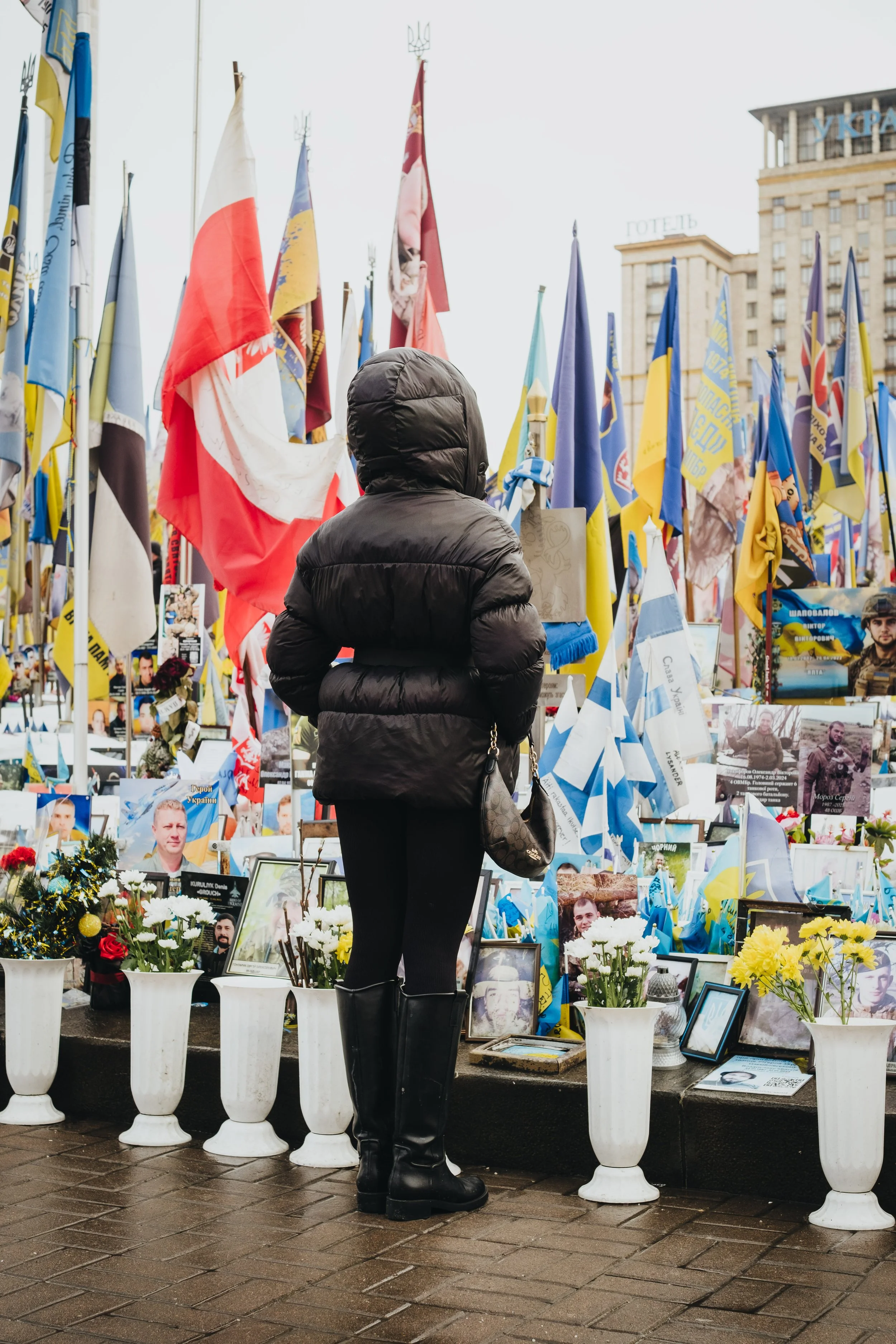 A person wearing a black puffy jacket and tall black boots standing in front of a memorial with flowers and photographs, surrounded by numerous flags.
