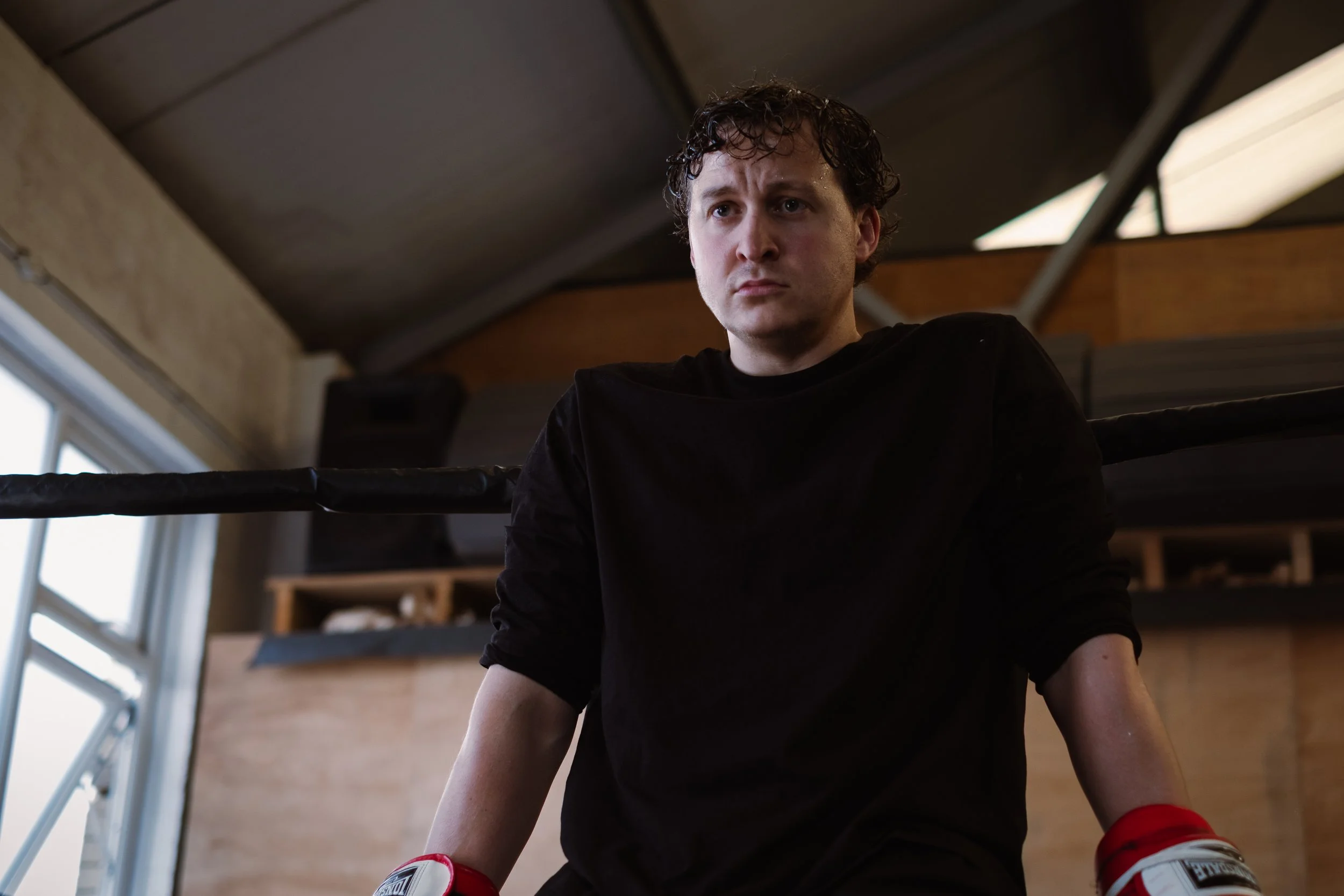 A young man with curly dark hair wearing a black shirt, sitting on a boxing ring, looking thoughtfully. He is inside a room with wooden ceiling, large windows, and shelves in the background. Red boxing gloves are on his lap.