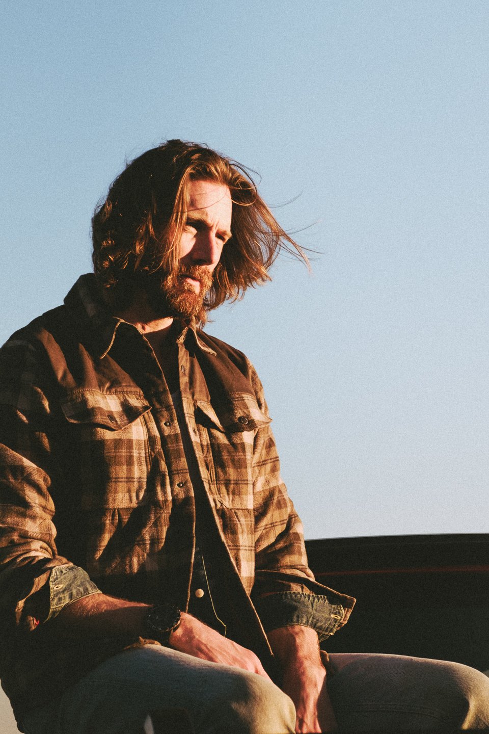A man with long hair and a beard wearing a plaid shirt, sitting outdoors during sunset or sunrise, with a clear blue sky in the background.