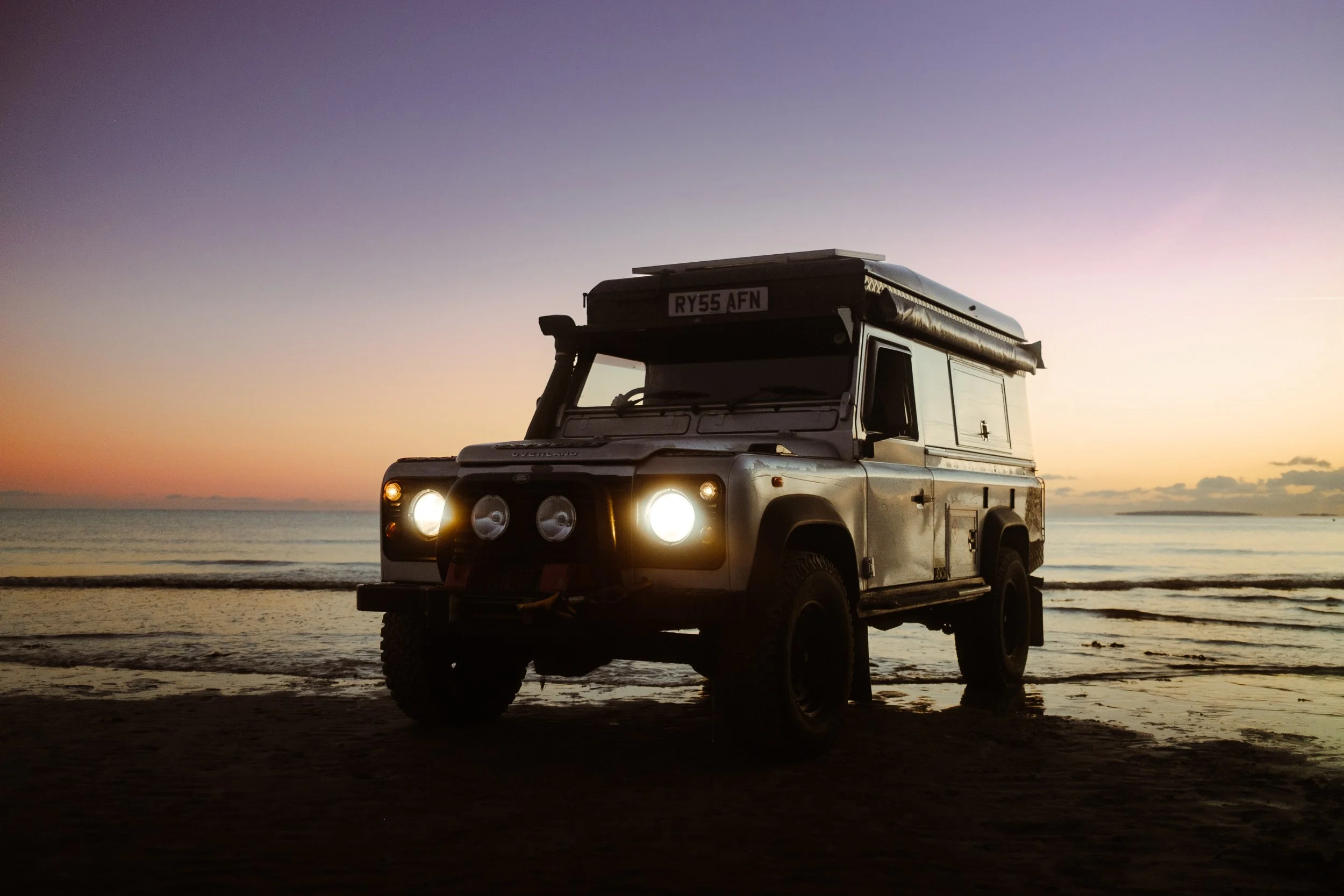A Land Rover Defender parked on a beach at sunset with the ocean in the background.