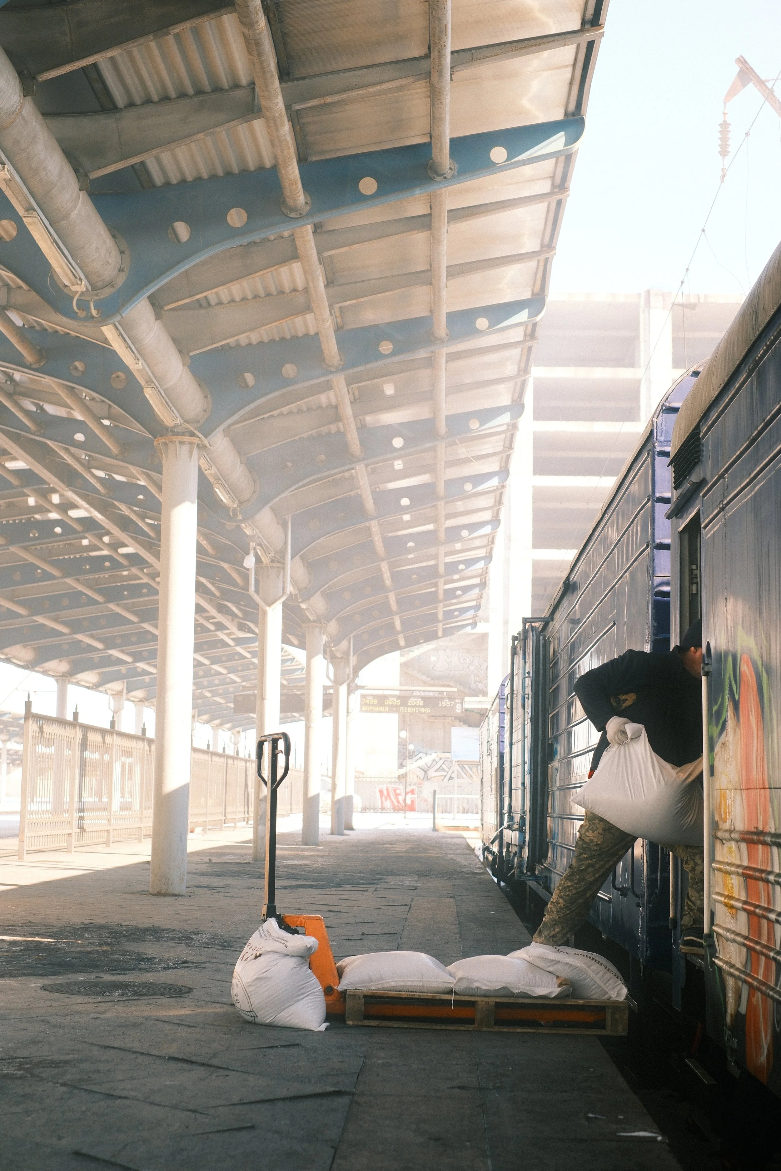 A train station platform with a covered roof structure. A person is partially visible, leaning out of a train with a door open. There are large bags and a wooden pallet on the ground, and a small barrier or sign is placed nearby. The area appears rel