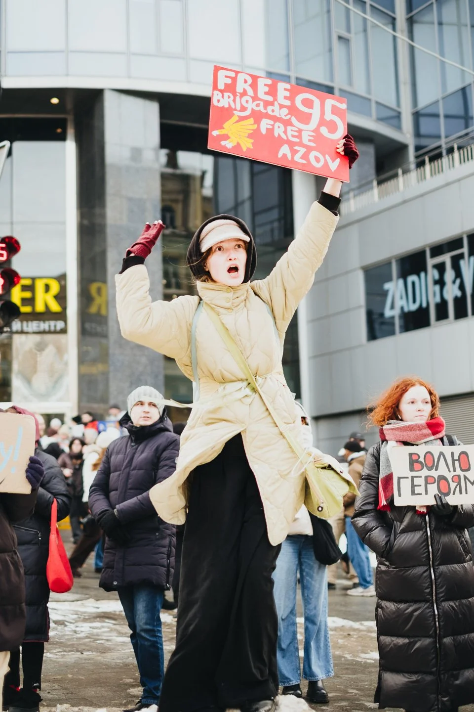 A woman protesting in a crowd, holding a red sign with white and yellow writing, including the number 95. She appears to be shouting or chanting, with other people around her holding signs, in an urban setting with tall buildings.