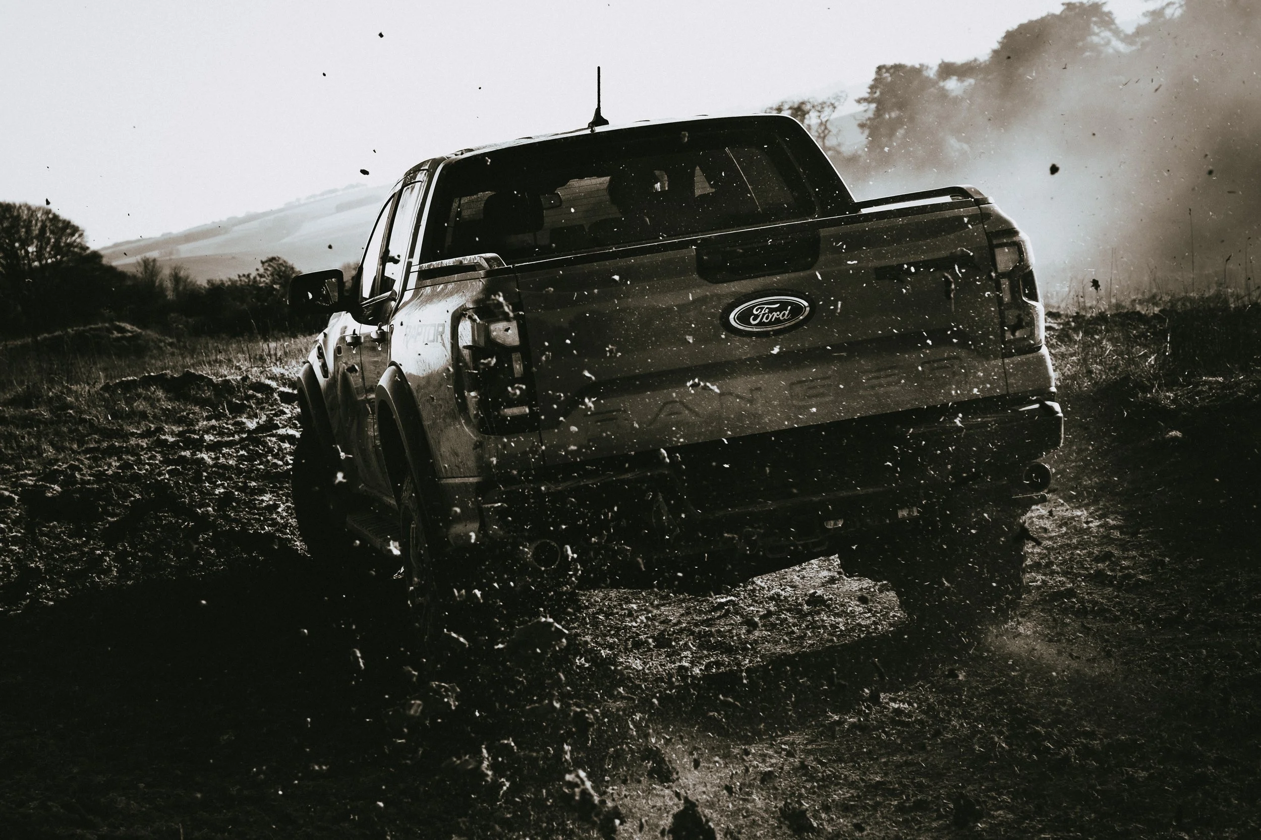 A black and white photo of a Ford pickup truck driving on a muddy off-road trail, kicking up dirt and debris, with trees and hills in the background.