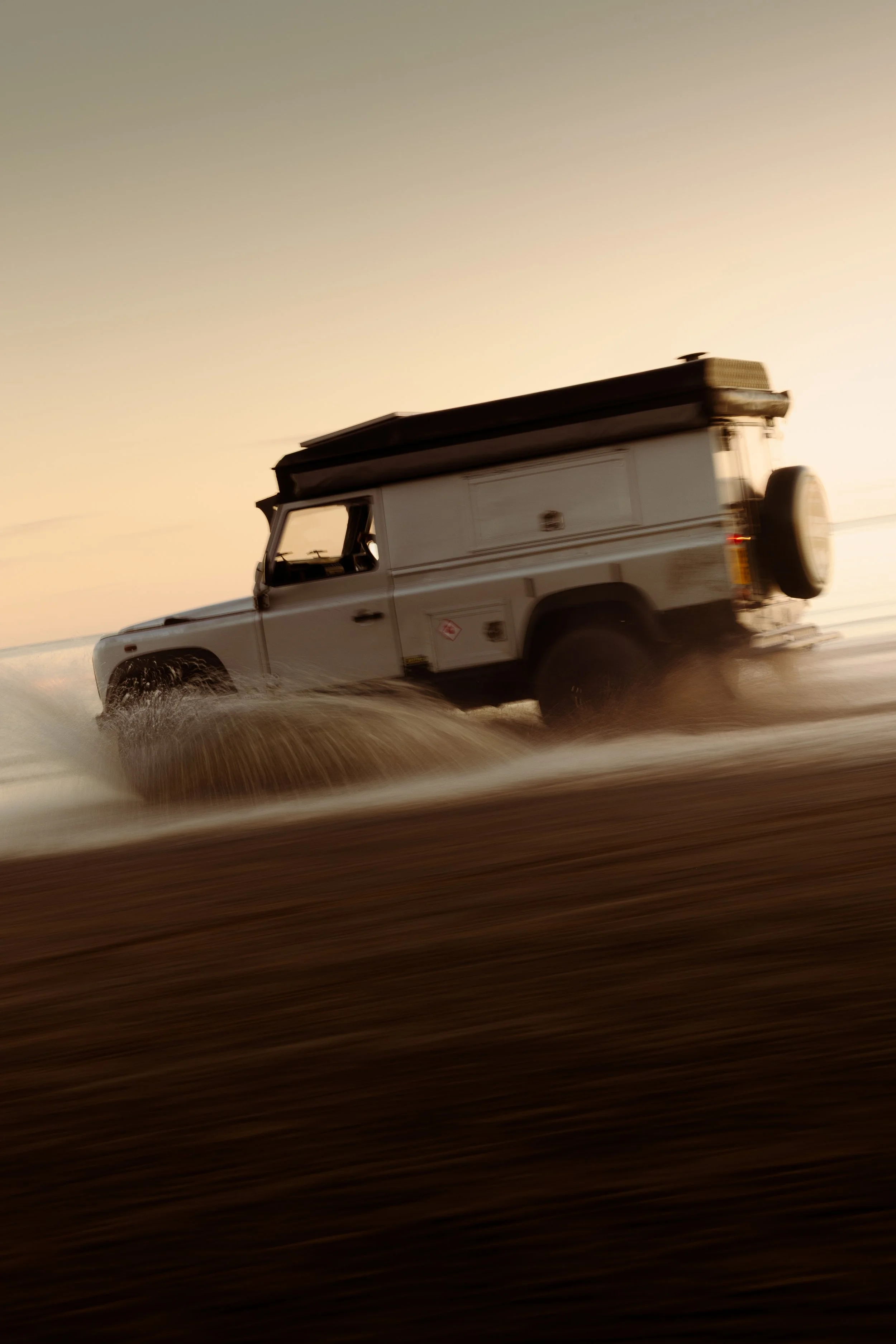 An off-road vehicle driving on a sandy beach at sunset, splashing water and sand as it moves.