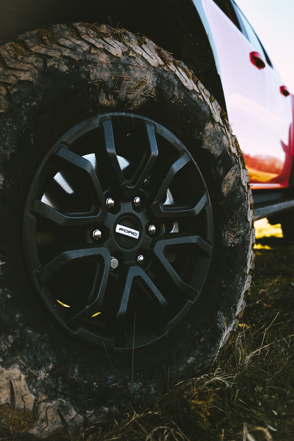 Close-up of a black Ford wheel with an off-road tire mounted on a muddy terrain, with part of a red vehicle body visible in the background.
