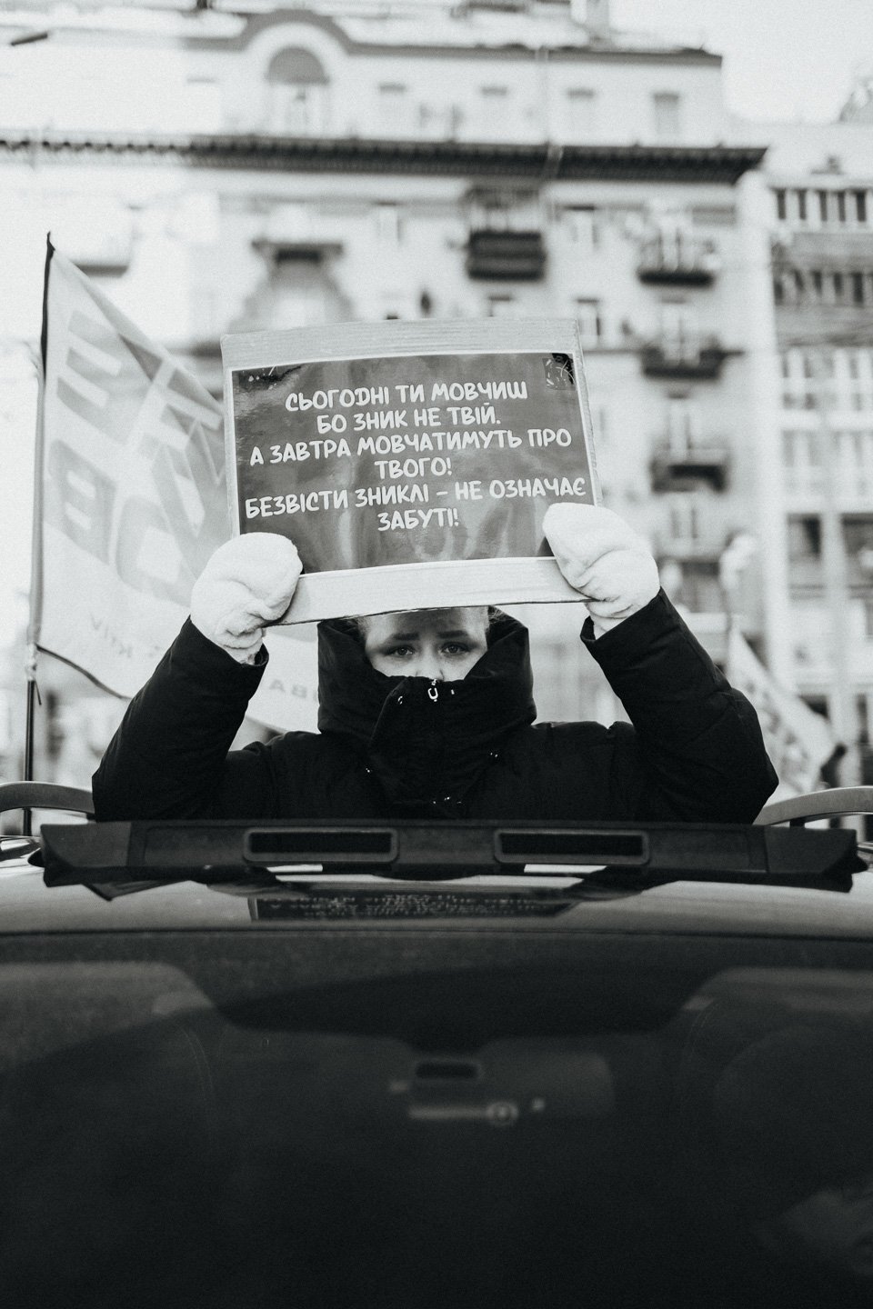 A person standing in front of a building, holding up a sign written in a language that appears to be Ukrainian, with a flag visible in the background.
