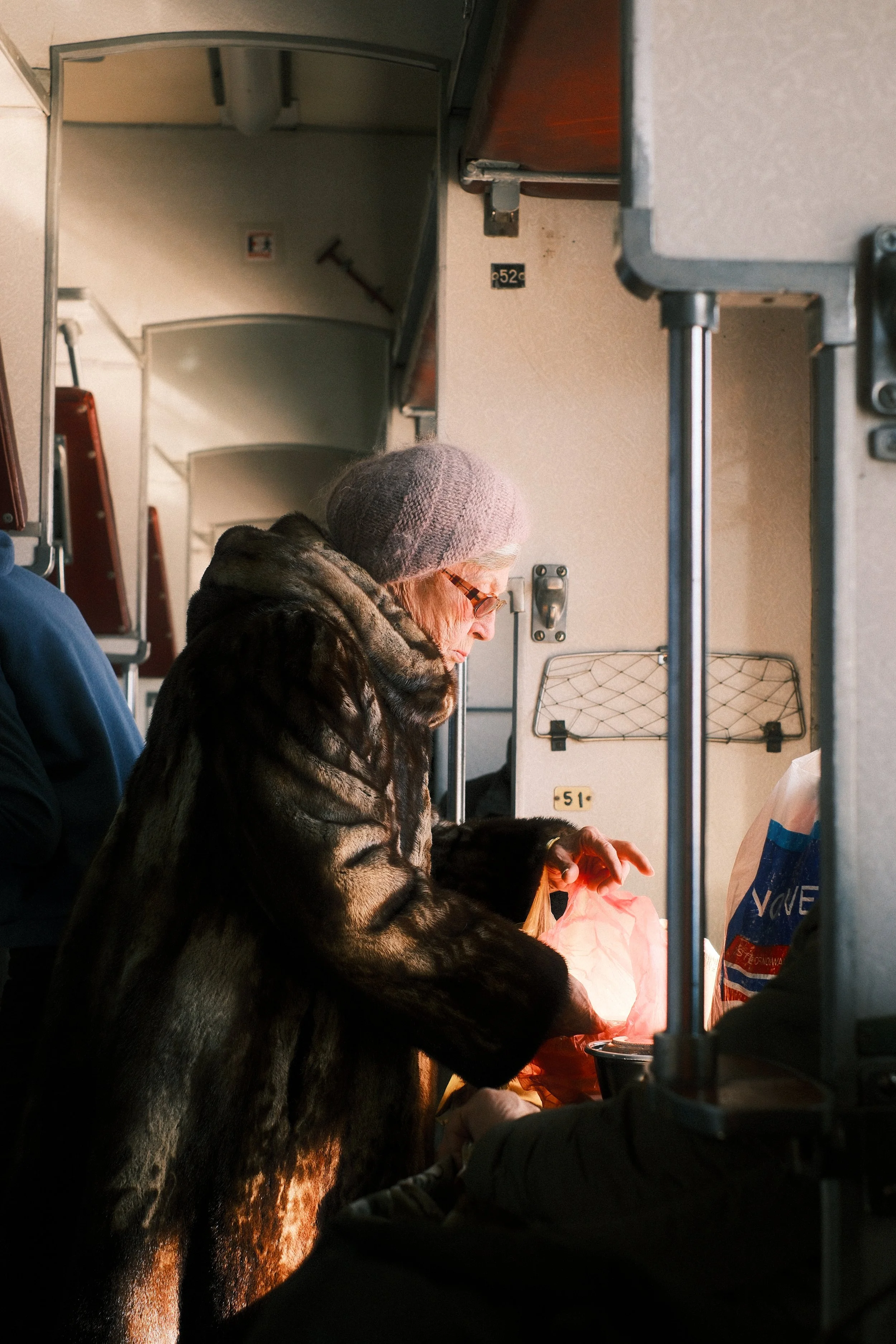 An elderly woman with glasses and a gray knit hat is in a bus, looking at a glowing bag or object, with a fur coat. Other passengers are visible, and the bus interior features overhead compartments and a handrail.