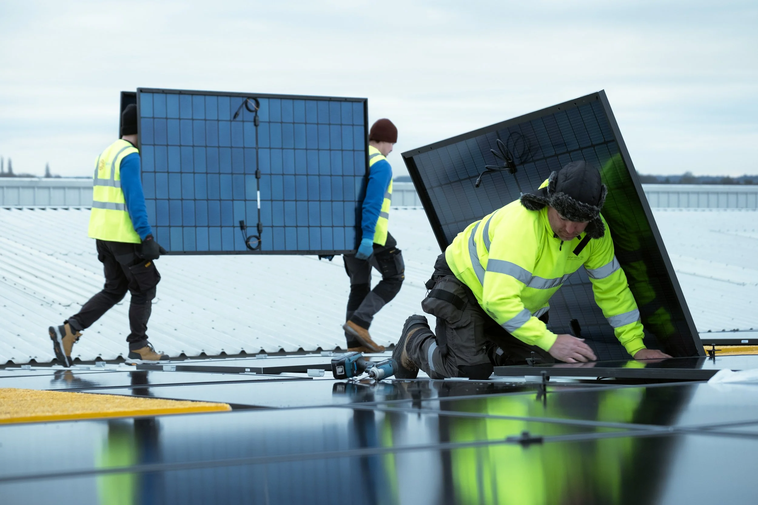Workers installing solar panels on a rooftop. One worker is kneeling, handling a solar panel, while two others carry additional panels into place.