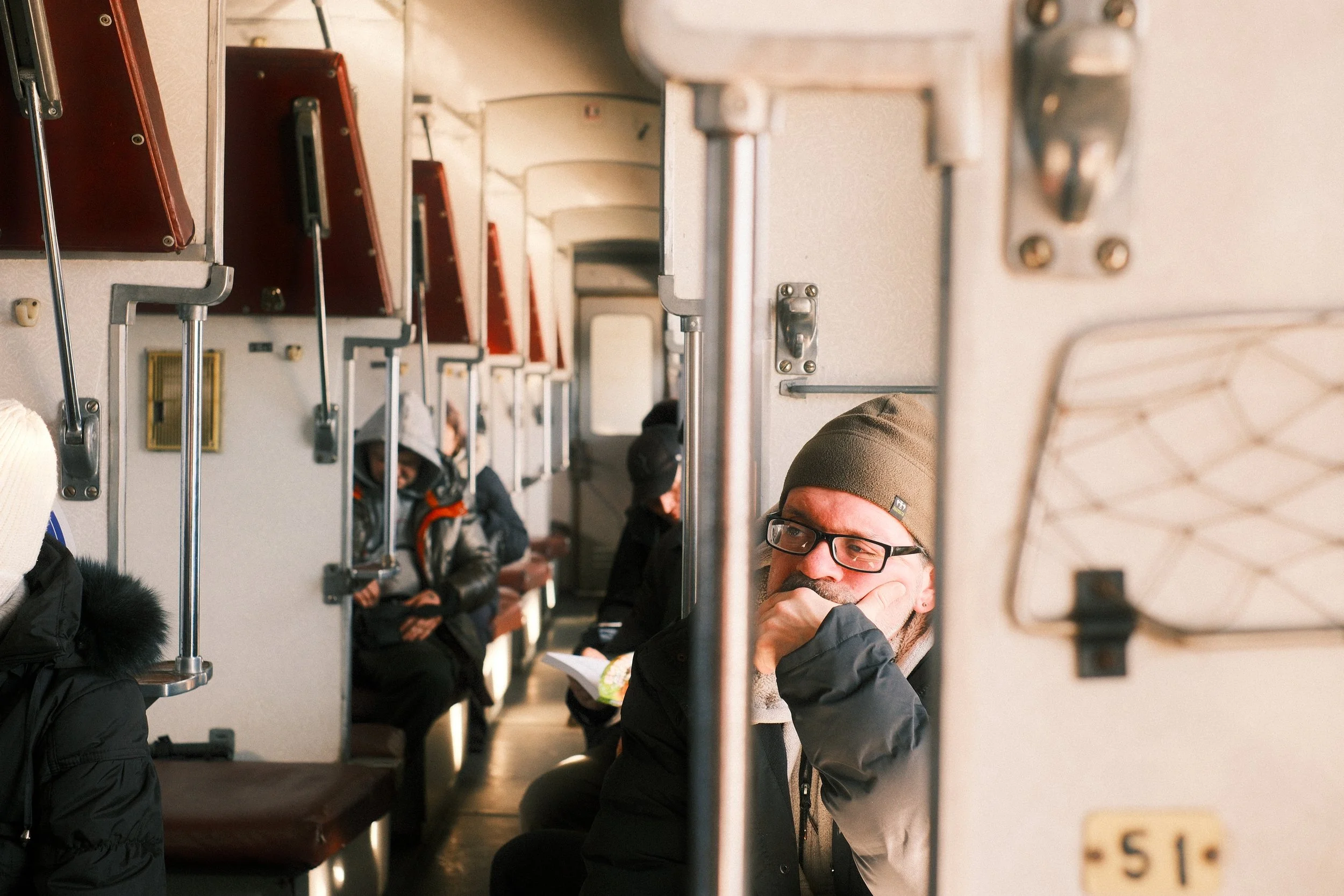 People sitting on benches inside a train. A man in a black jacket and glasses is resting his chin on his hand, looking thoughtful. Other passengers are reading or looking down.