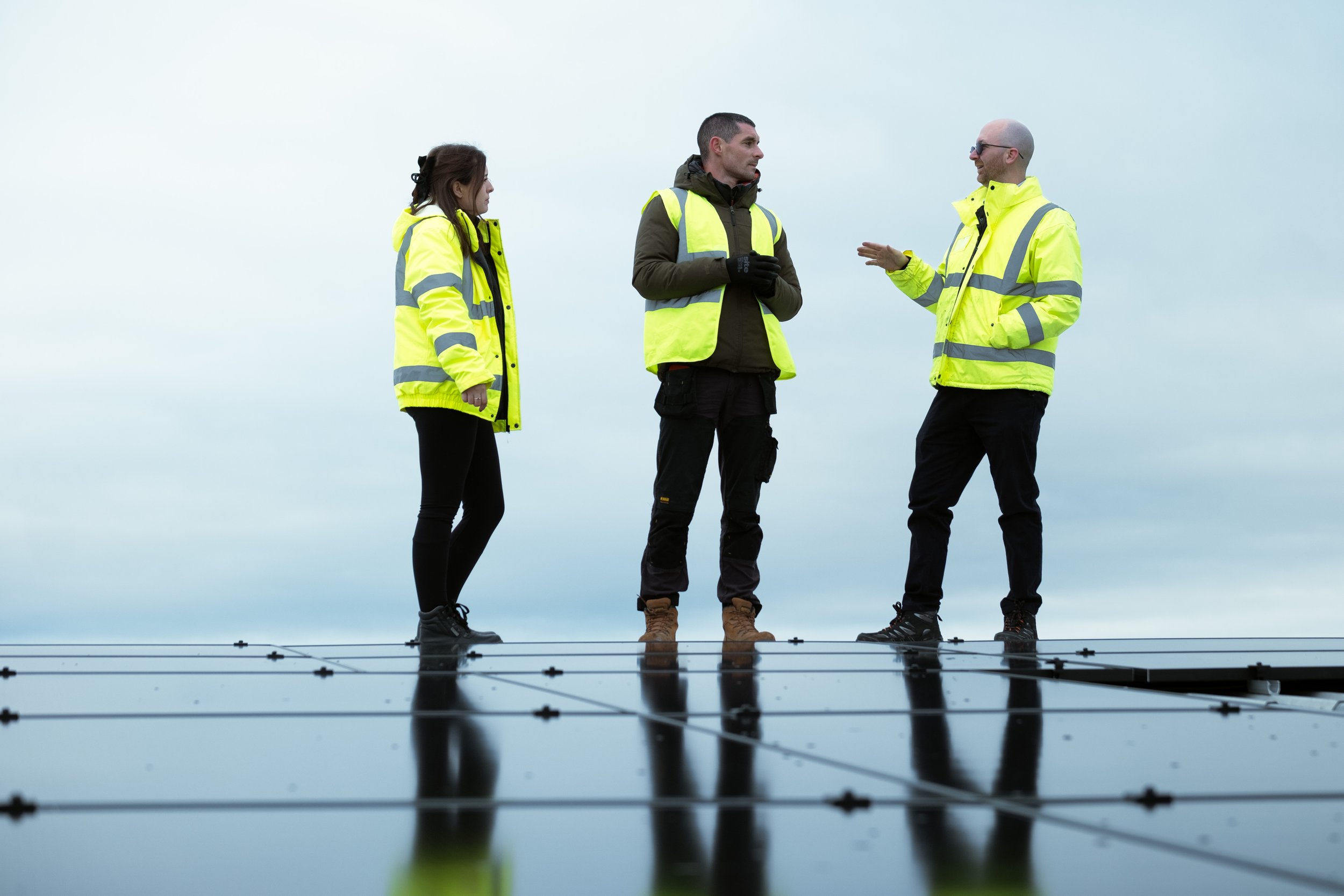 Three people in high-visibility jackets standing on a solar panel. They are talking, with one person gesturing with his hand.