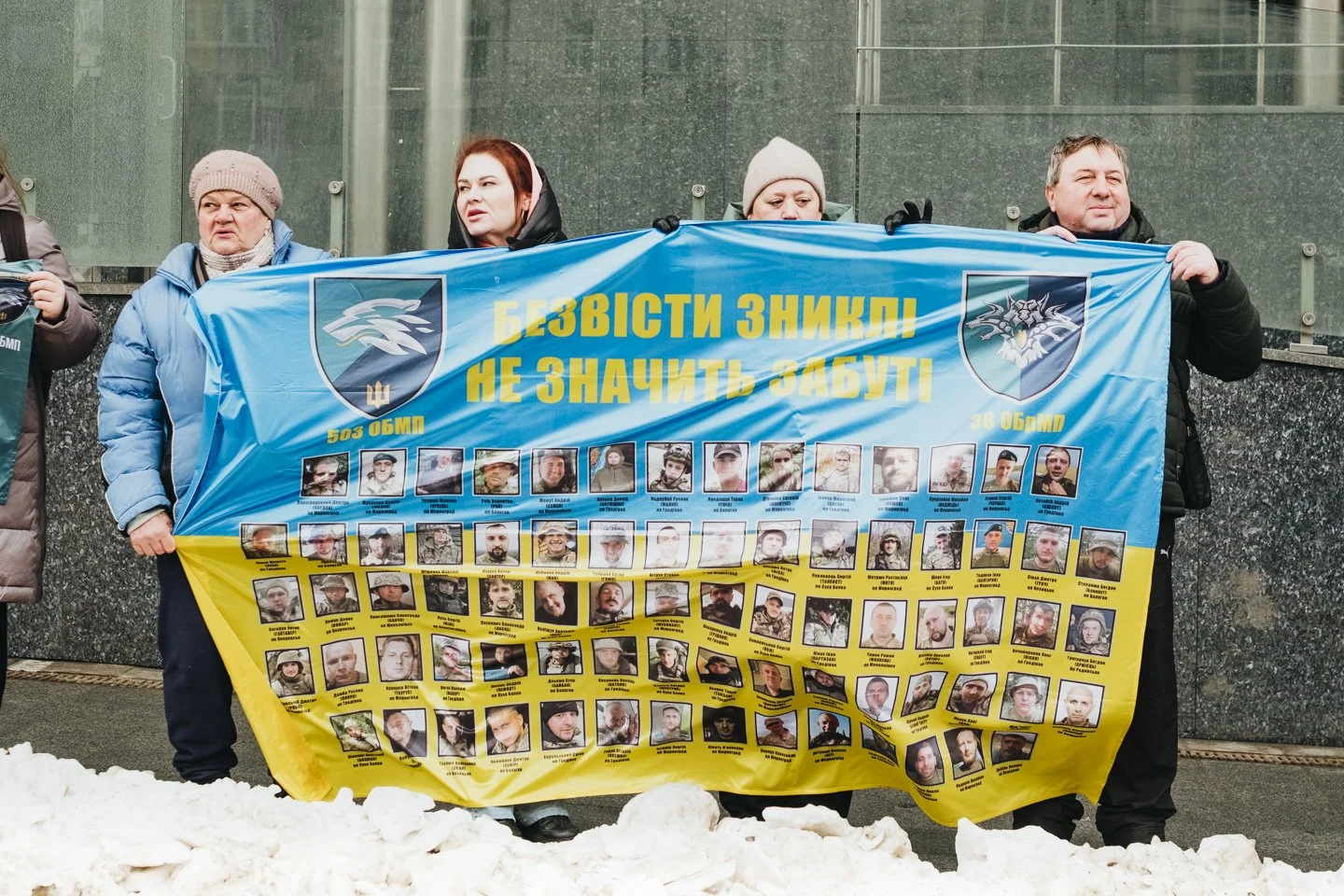 People holding a large banner with photos of missing persons and Ukrainian text, standing in front of a building with snow on the ground.