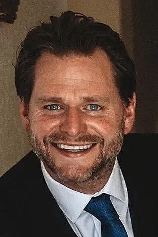 A smiling man with dark hair and a beard, wearing a suit and tie.