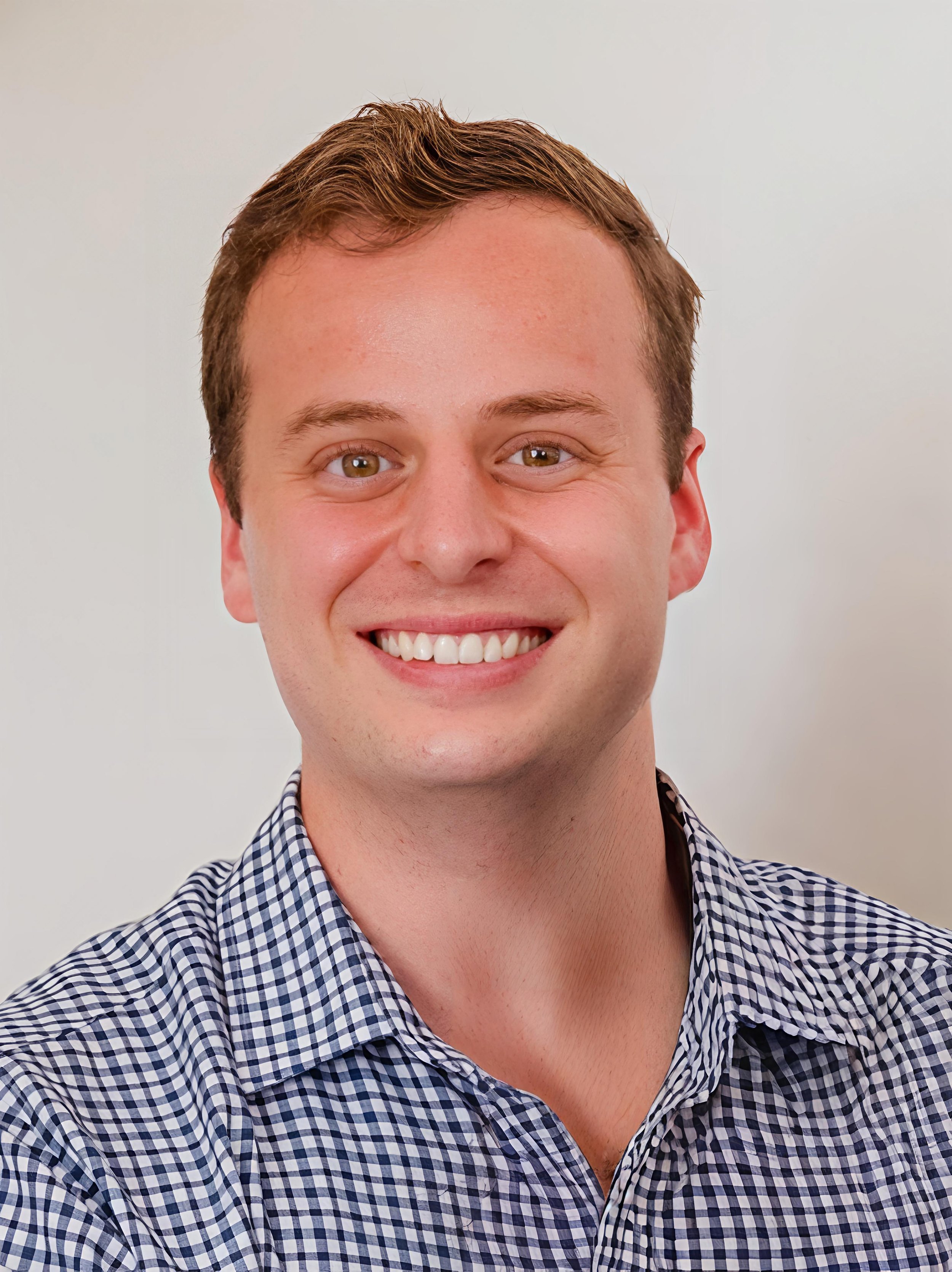 A smiling young man with light skin, brown hair, and brown eyes wearing a checkered shirt.