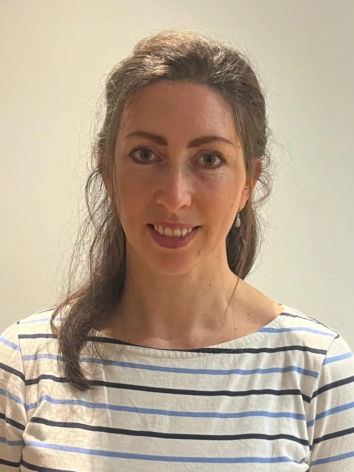 A woman with long brown hair, blue eyes, and a striped shirt smiling indoors against a light-colored wall.